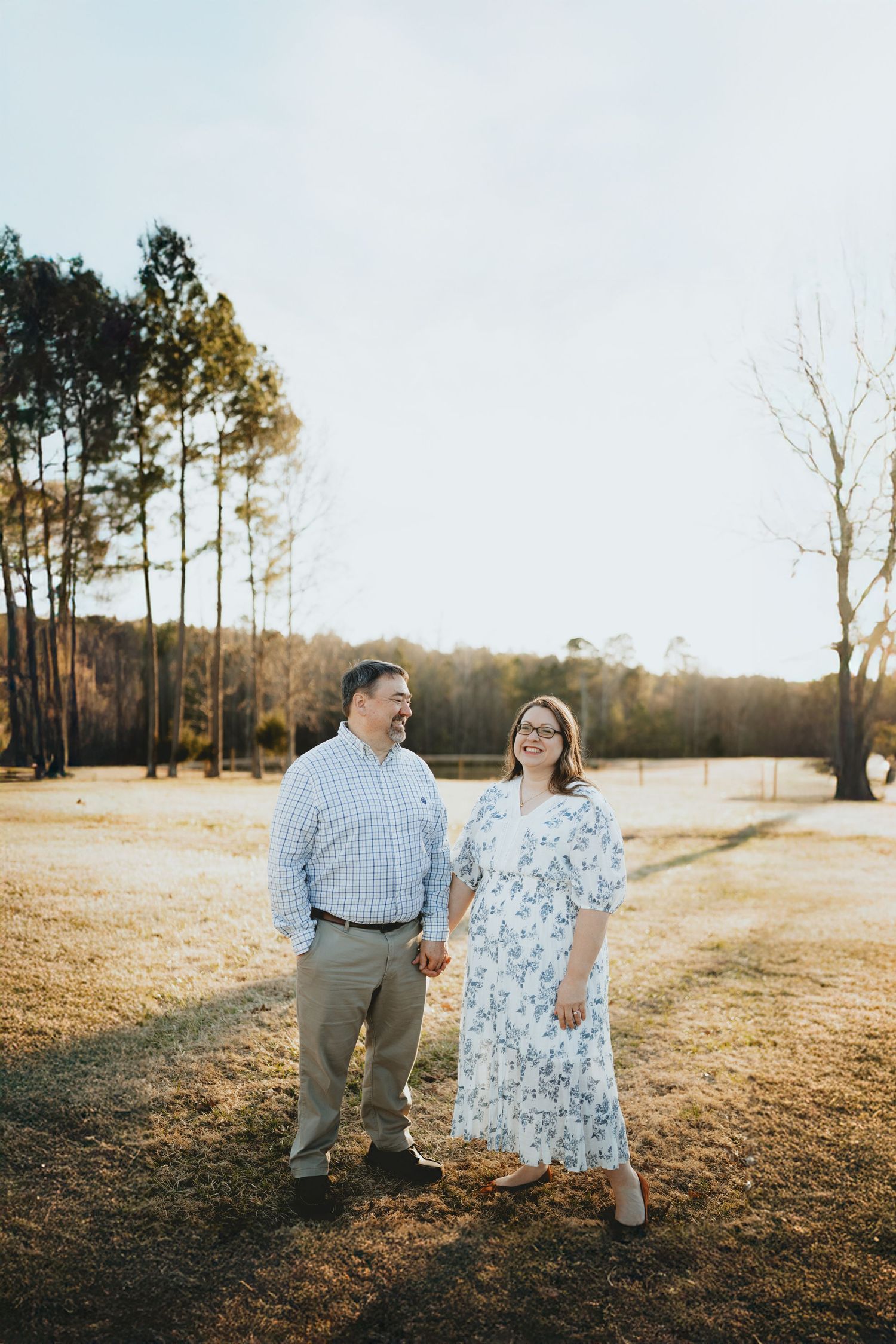 Smiling couple holding hands and walking together in a golden field surrounded by tall trees.