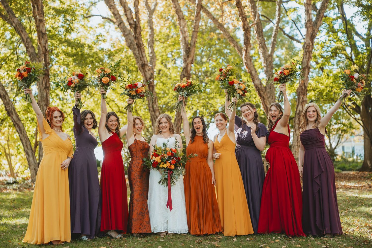 Bride with bridesmaids in mismatched fall-colored dresses raising bouquets under autumn trees.