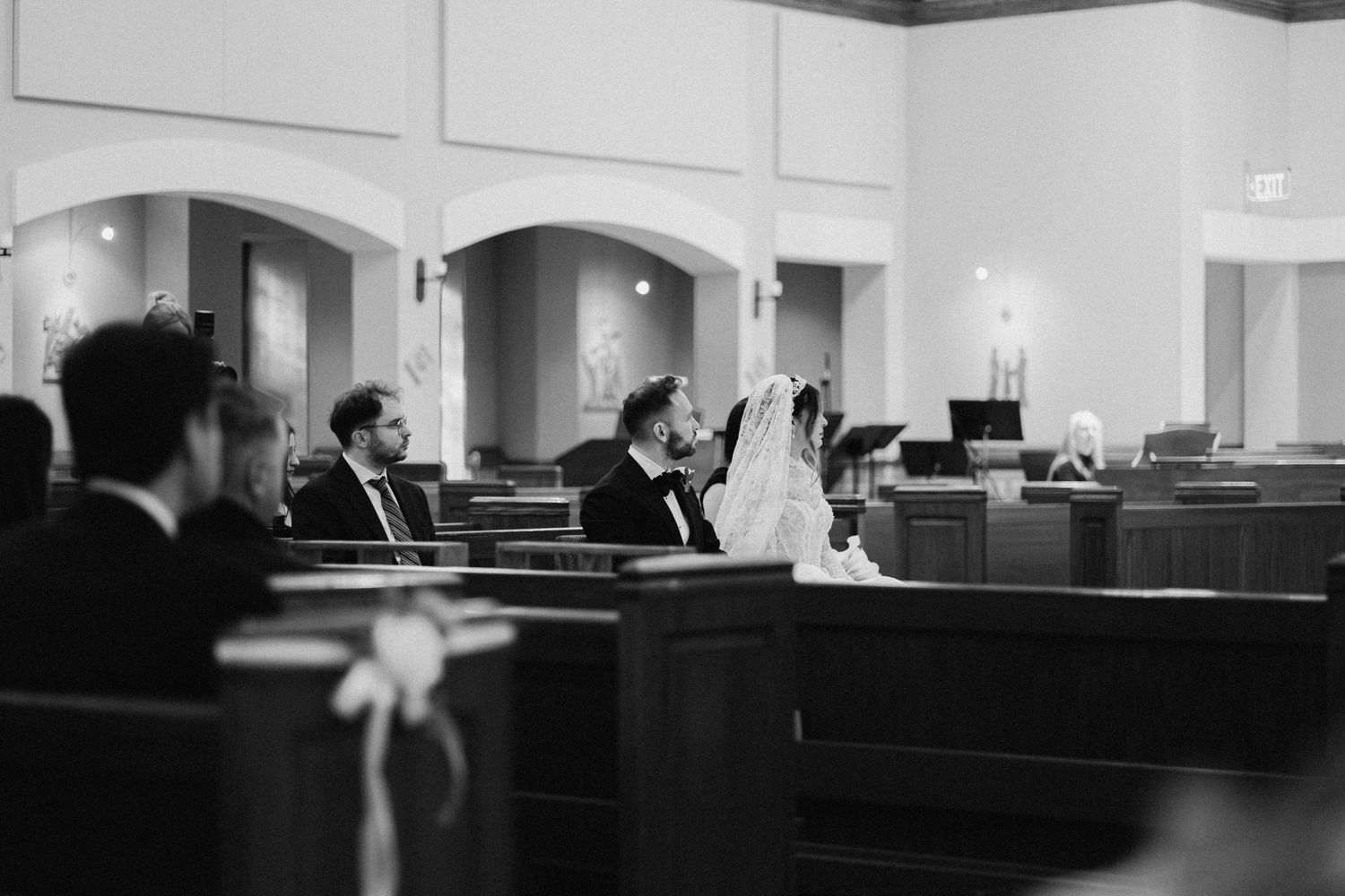 Black and white wide shot of bride and groom seated in church pew during ceremony.