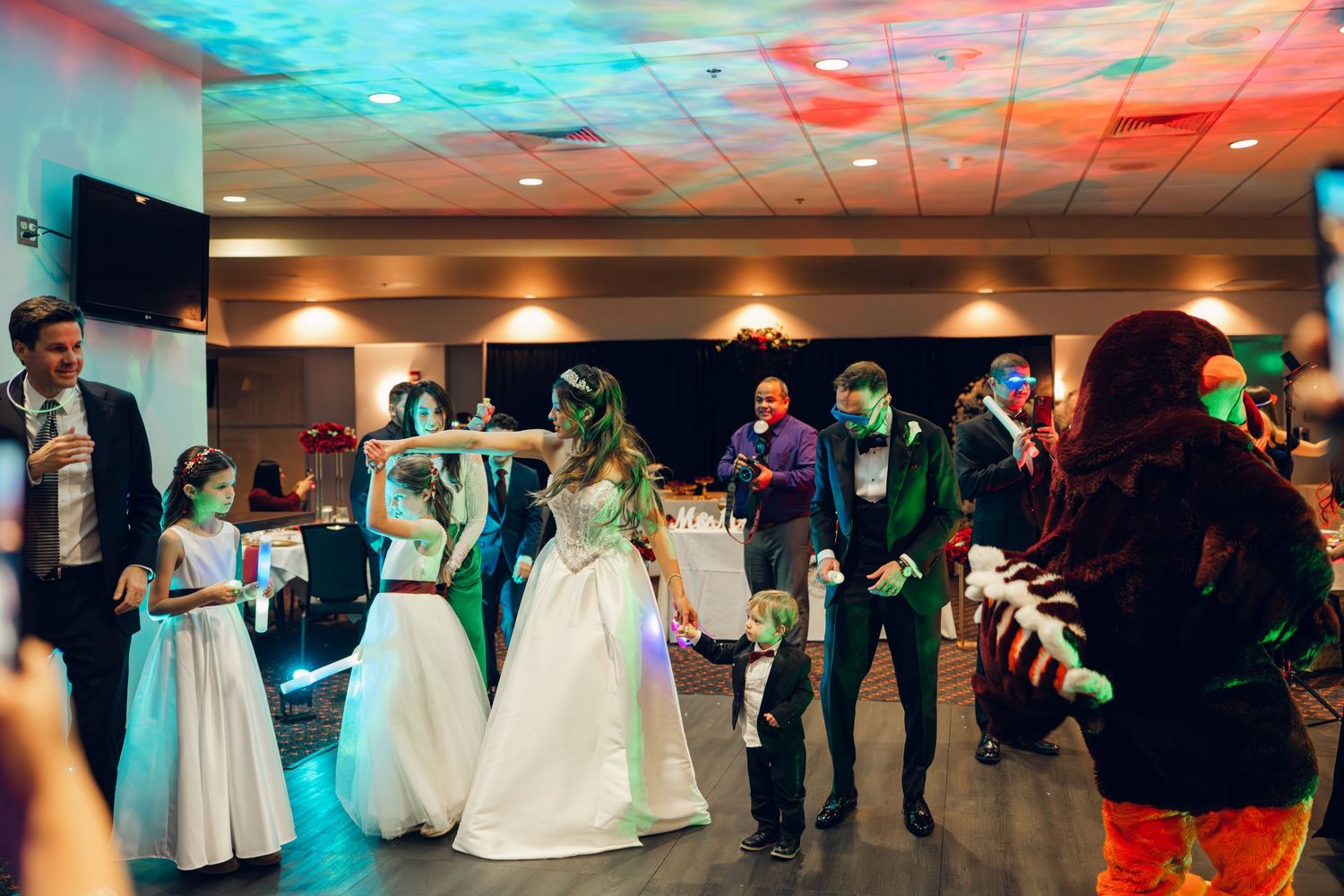 Bride and groom dance with guests under colorful lights at festive wedding reception.