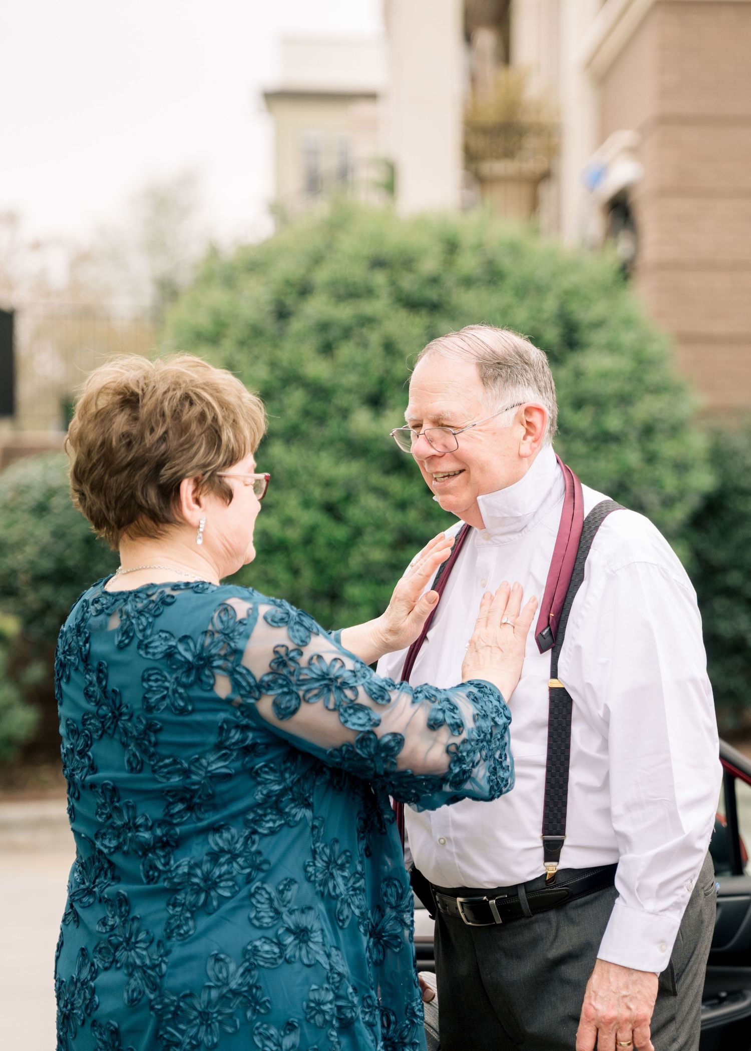 Elderly couple sharing a warm moment outdoors at a wedding, woman in teal lace dress touching mans chest.