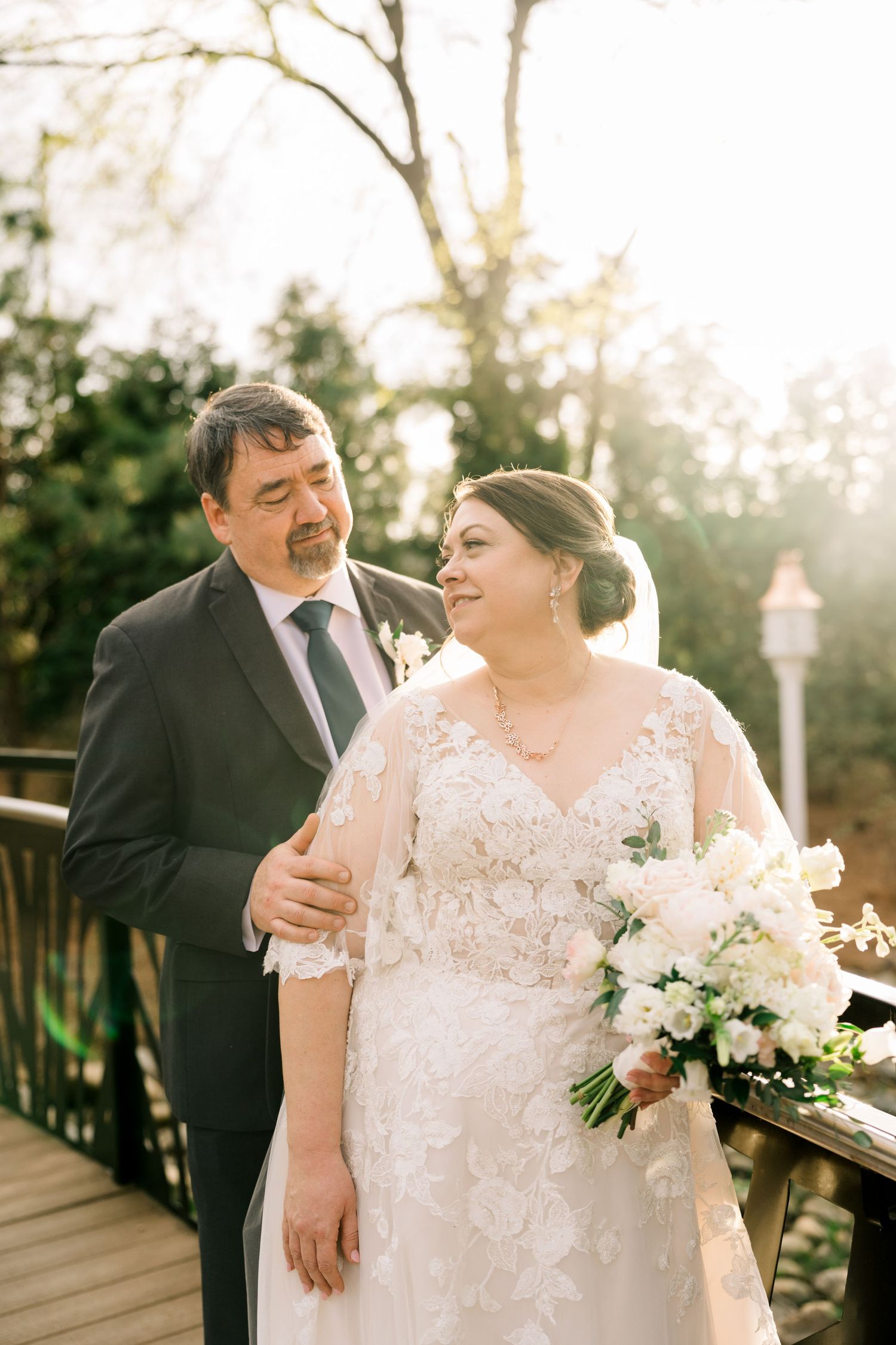 Bride and groom sharing a tender moment on a sunlit bridge, bride holding white floral bouquet.