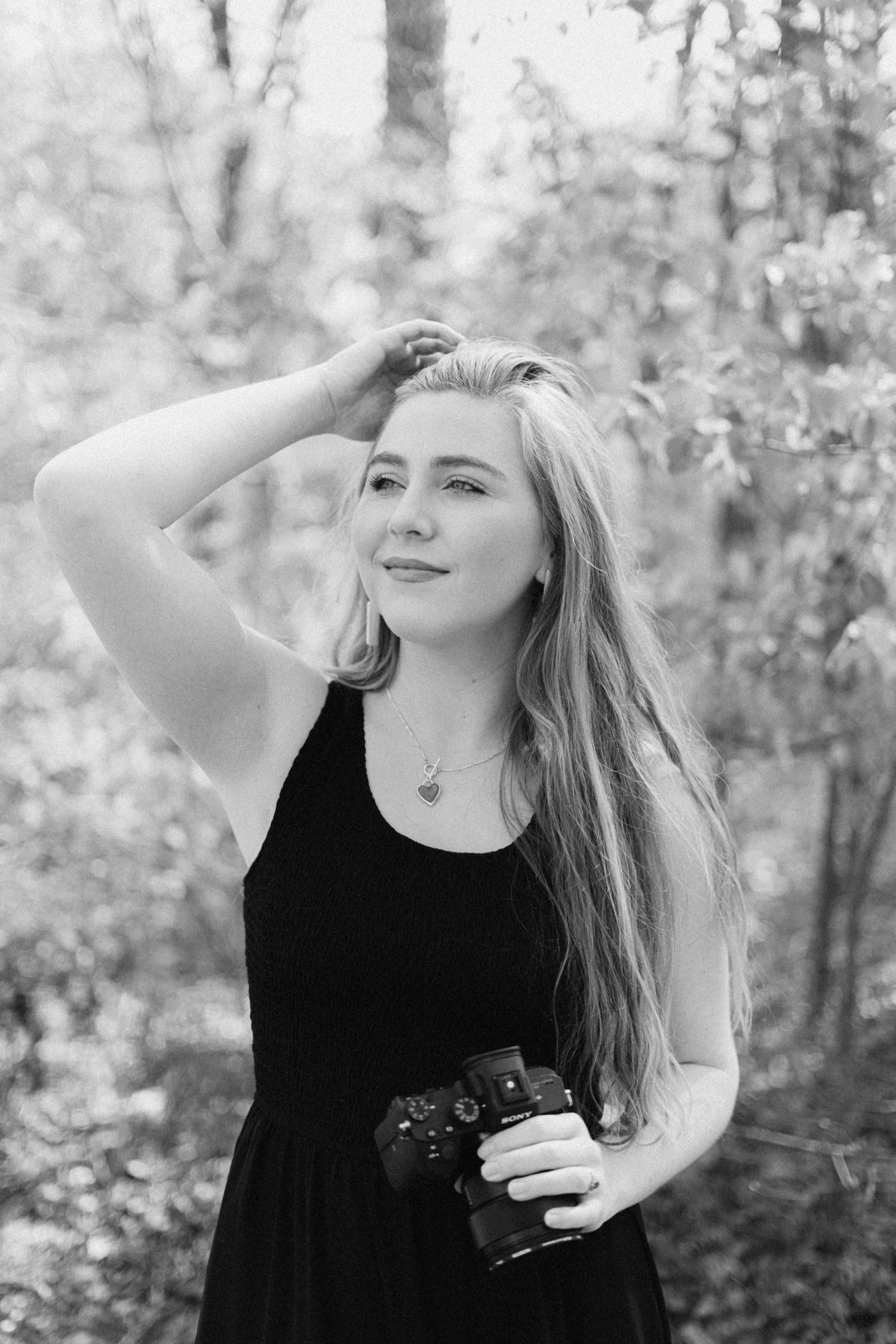 Blonde woman with DSLR camera in woodland setting, wearing black dress, hand raised to hair, monochrome.