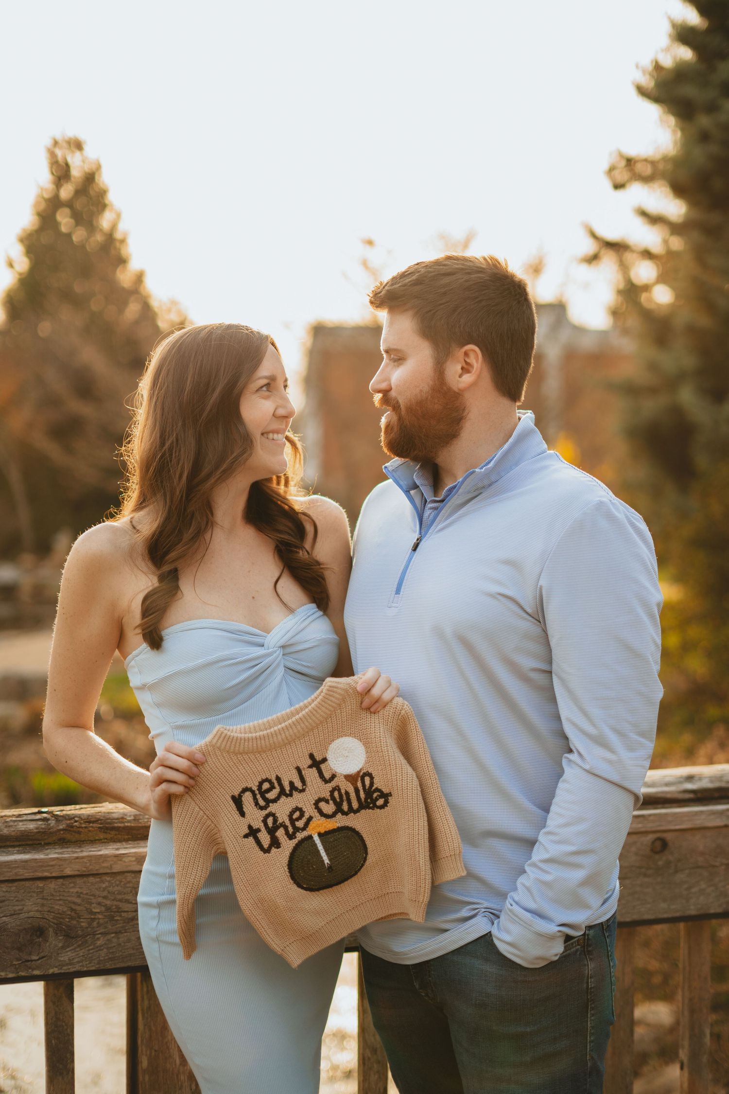 Couple holding a baby onesie that reads 'new to the club' during a golden hour maternity photo session outdoors.
