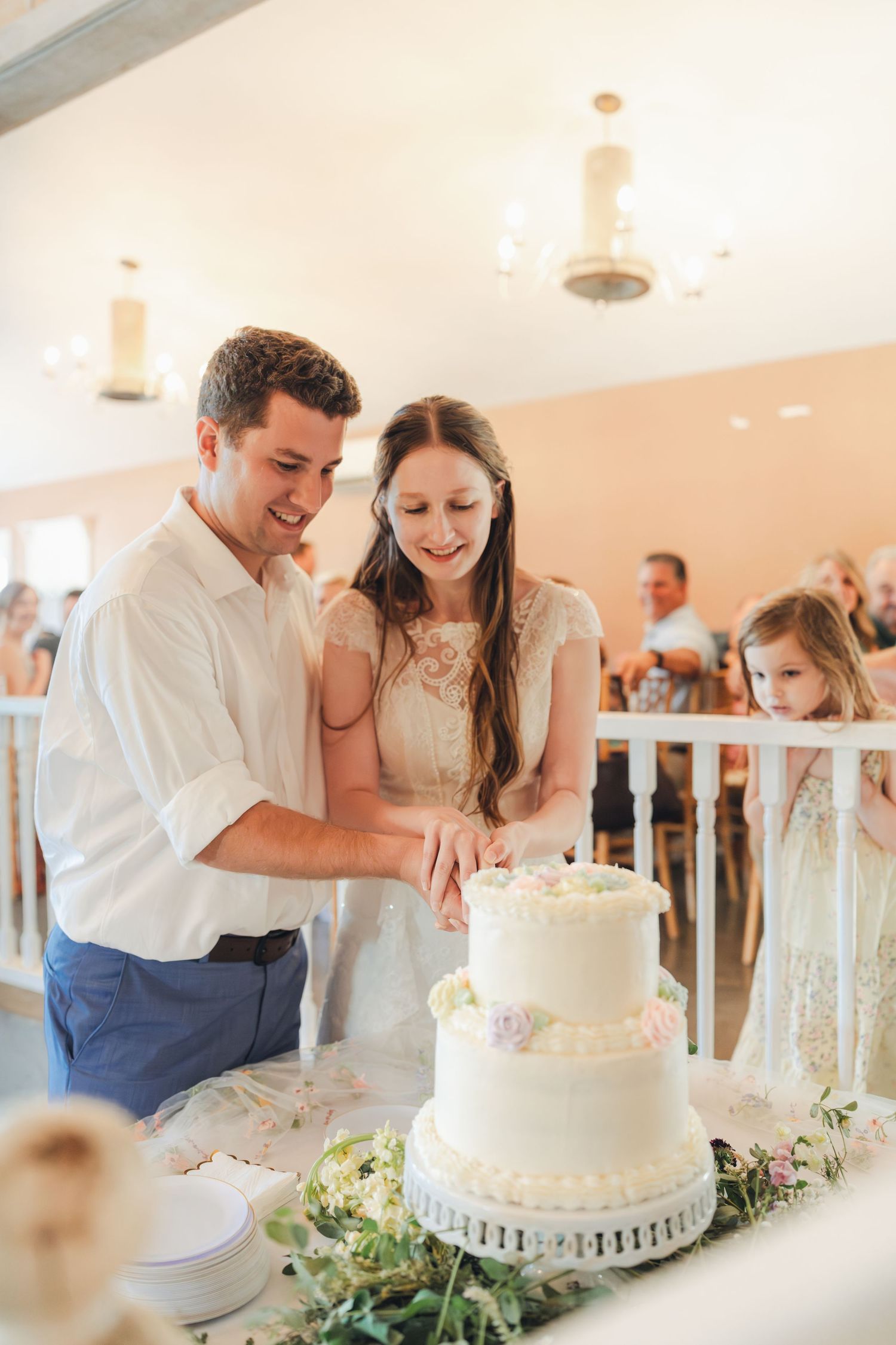 Smiling newlyweds cutting a three-tier white wedding cake together at their elegant indoor wedding reception.
