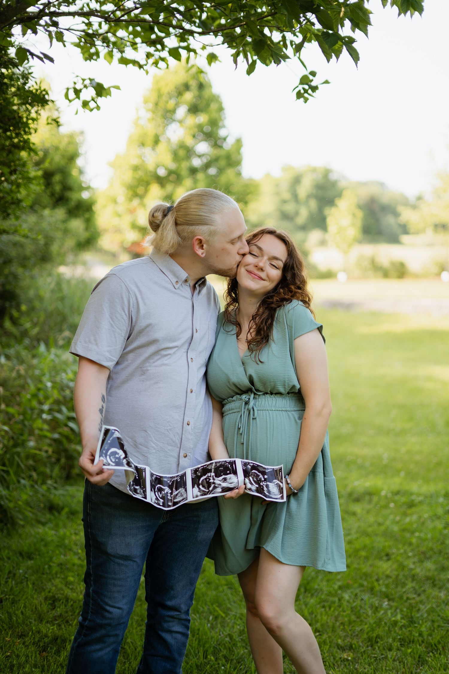 Couple sharing kiss while holding ultrasound photos outdoors under leafy tree in summer.