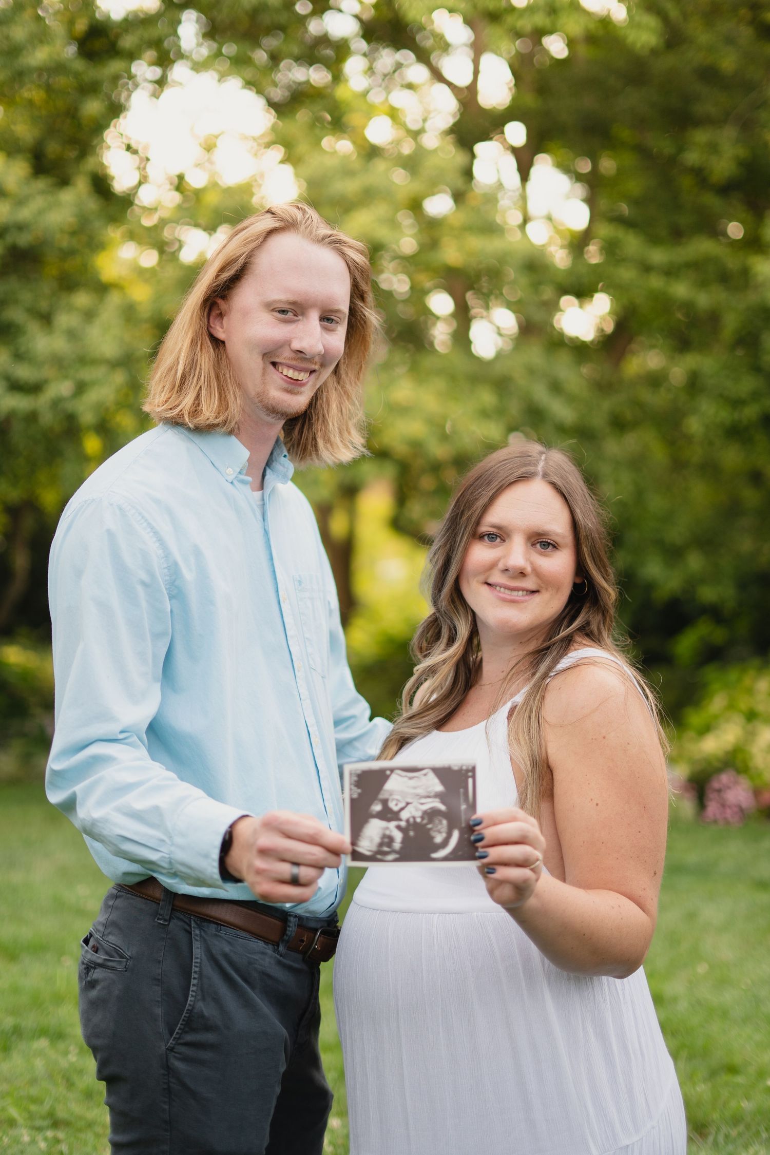 Smiling couple proudly displays ultrasound photo outdoors in a sunny green garden setting.