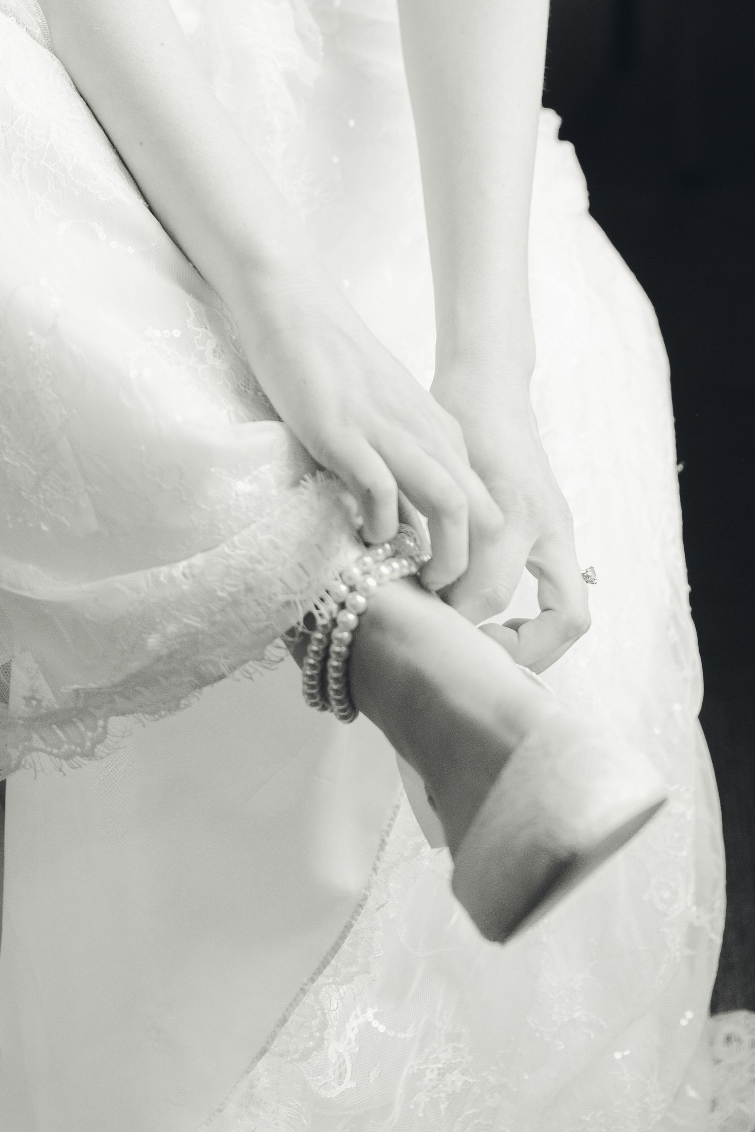 Black and white photo of a bride in lace dress fastening a pearl bracelet on her wrist.