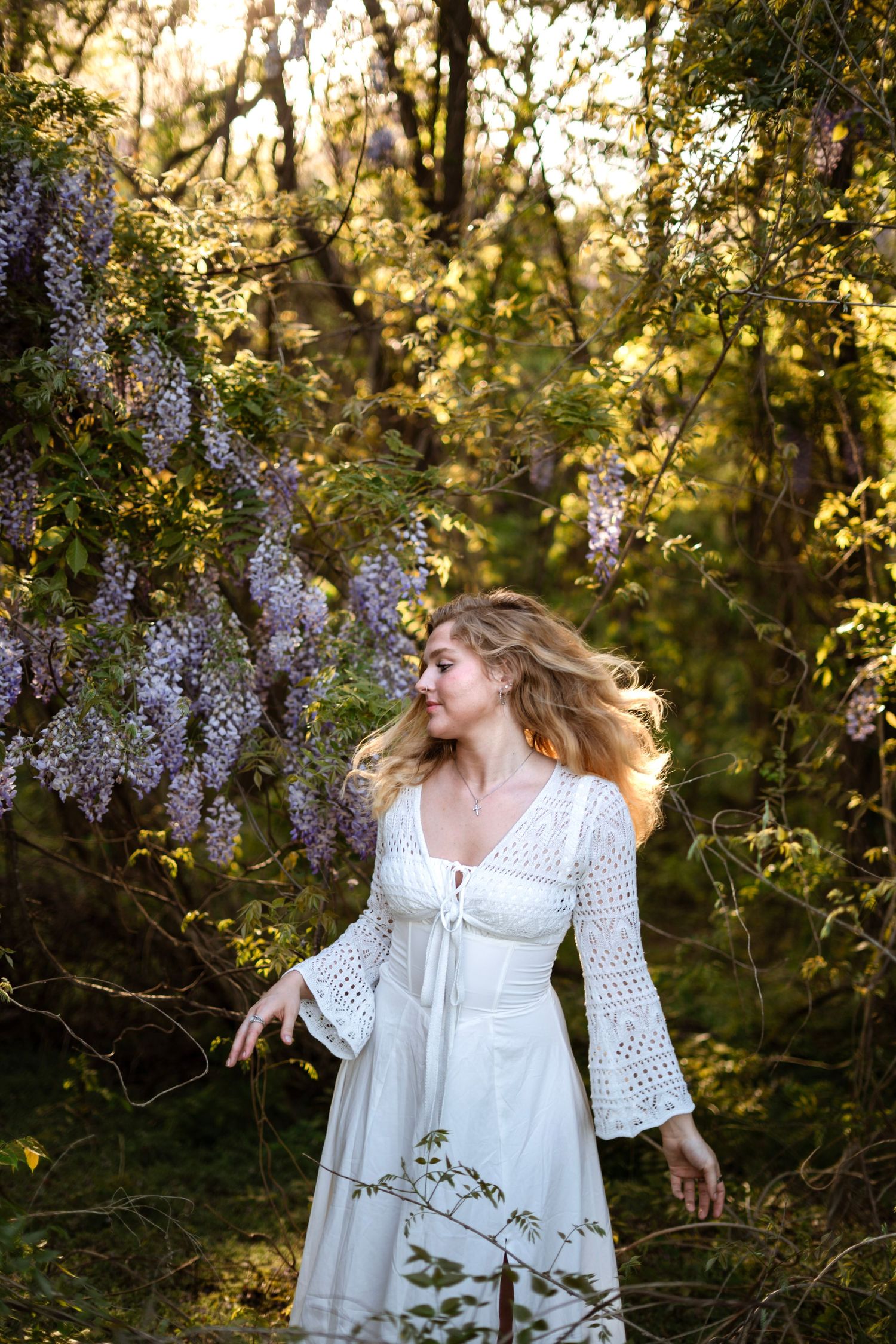 Blonde woman in white crochet dress stands among blooming purple wisteria in golden forest light.