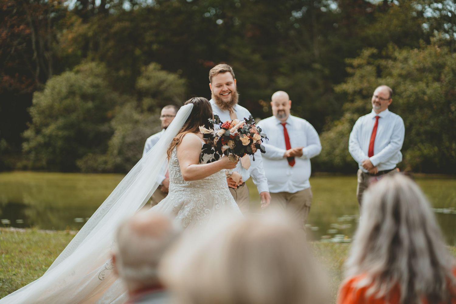 Smiling groom holds bouquet beside bride in lace dress and veil at outdoor wedding ceremony.