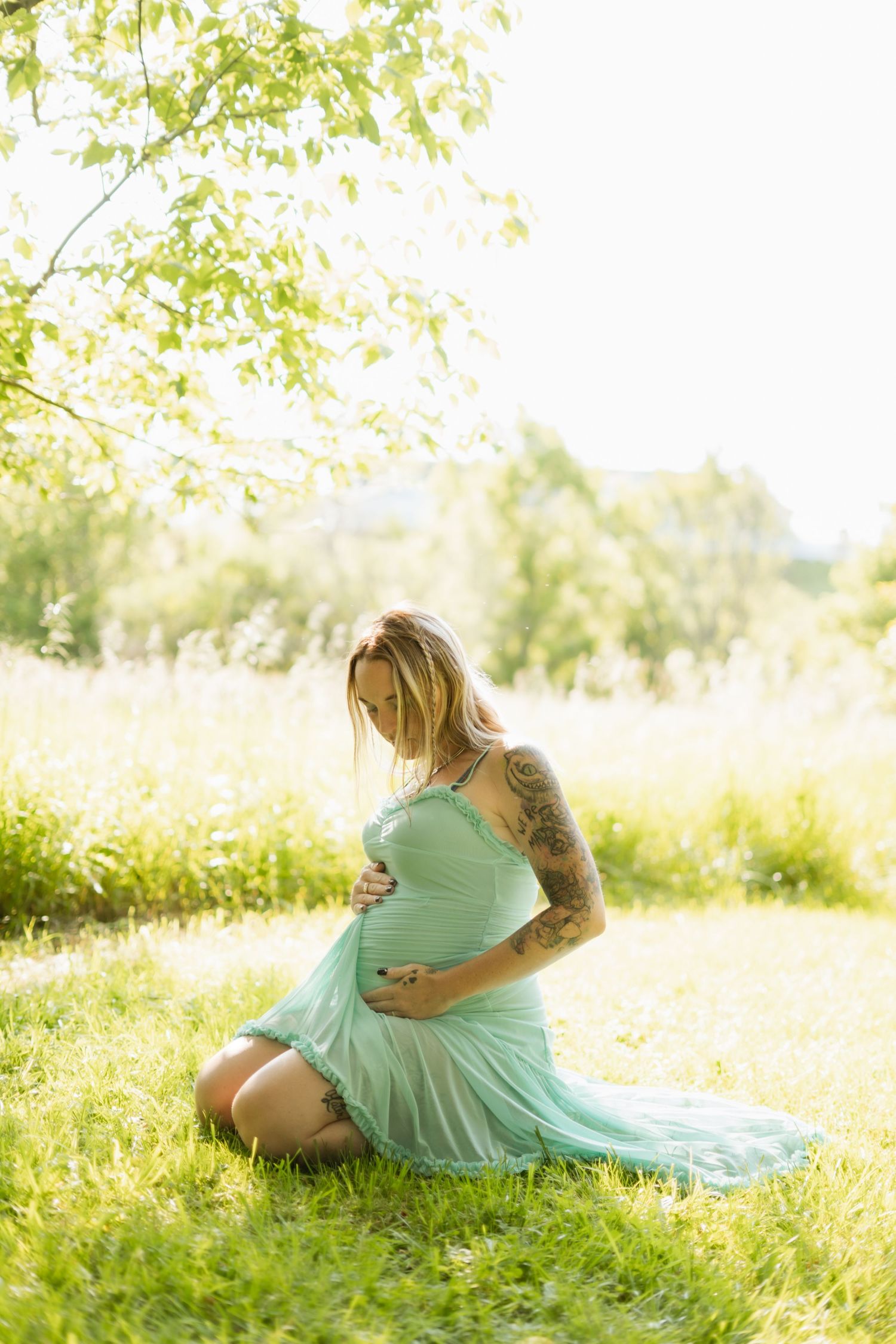 Tattooed pregnant woman in mint dress sitting on grass cradling baby bump in sunny outdoor setting.