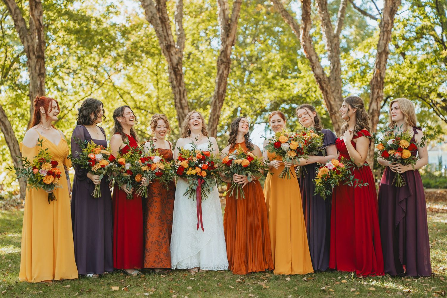 Bride surrounded by bridesmaids in mismatched fall-colored gowns holding vibrant autumn bouquets.