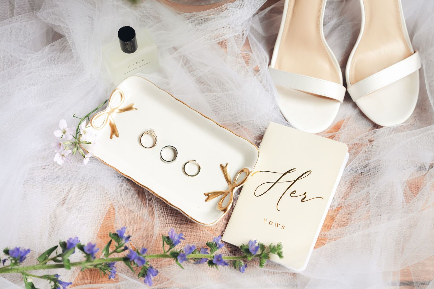 Wedding flat lay with rings, white heels, floral accents, perfume, and a gold-script Her vow book on soft fabric.