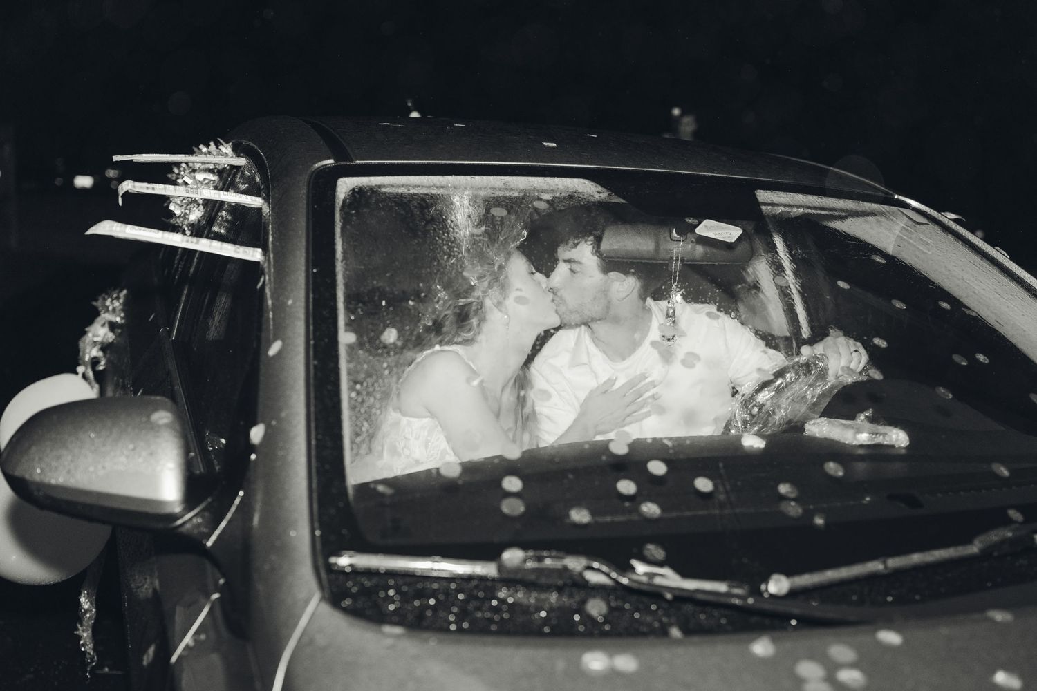 Couple sharing romantic kiss inside car captured in black and white wedding photo.