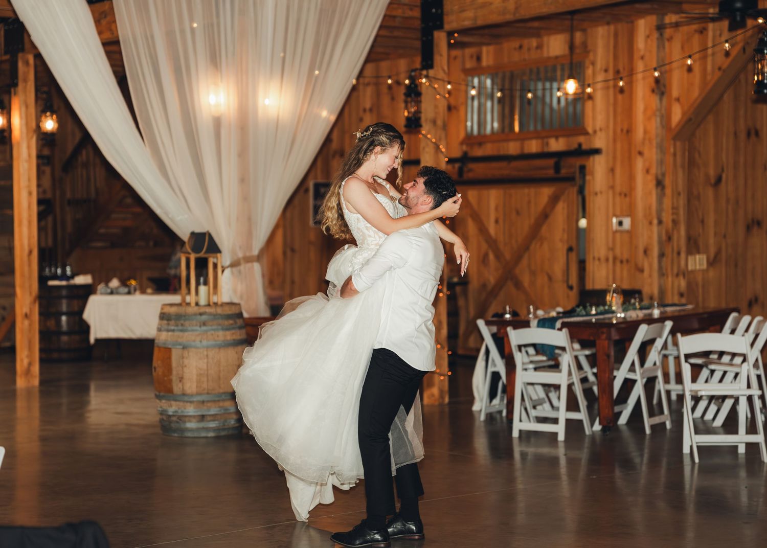 A groom lifts his bride during their first dance inside a rustic barn wedding reception venue.