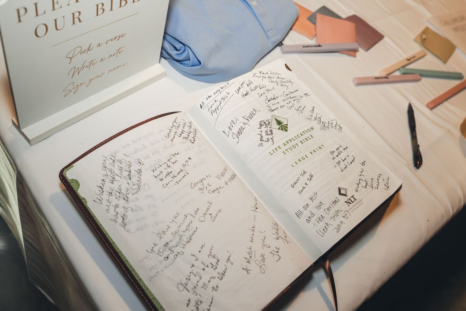 Open Bible guest book filled with handwritten wedding messages, surrounded by colorful pens on a white table.