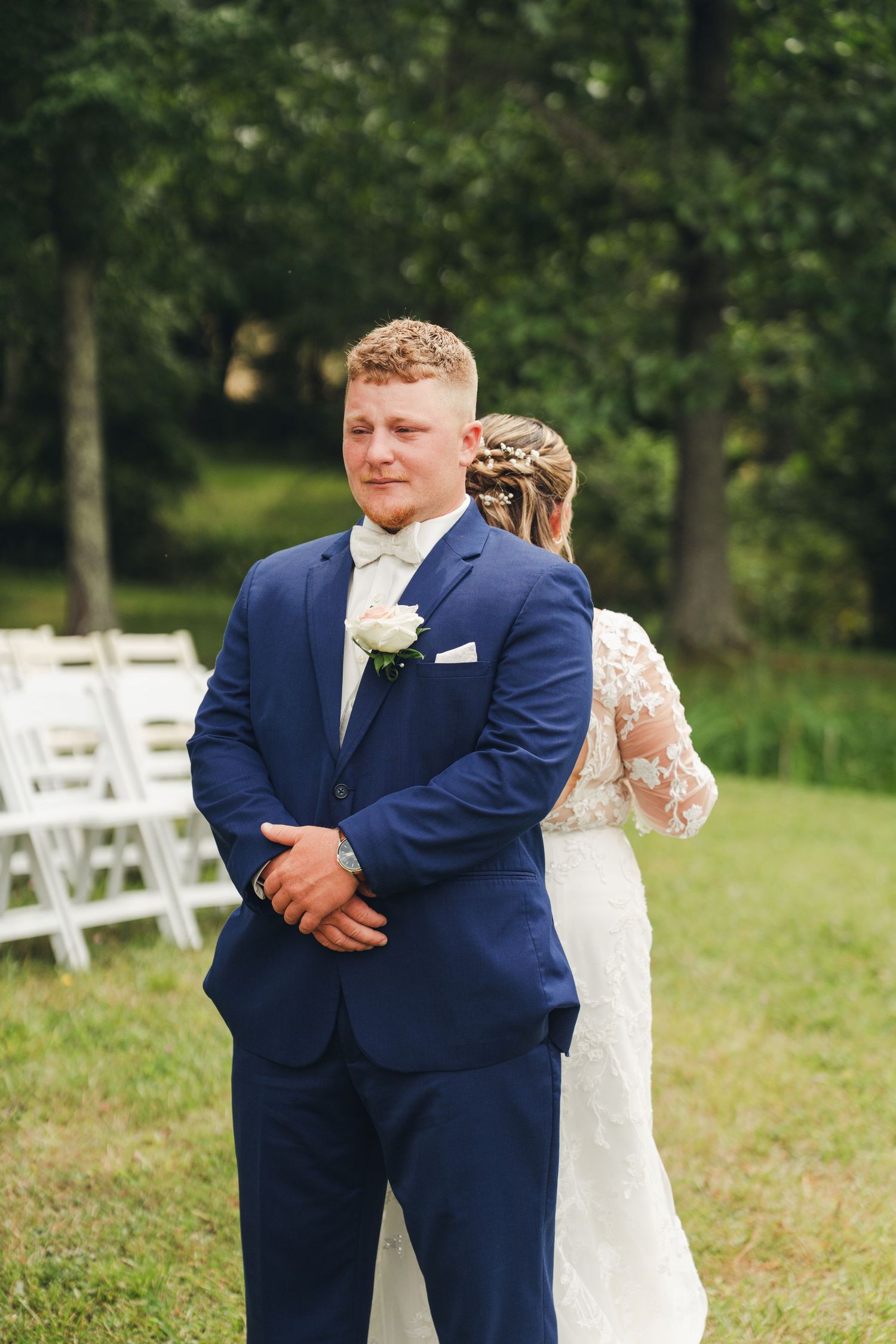 Groom in navy blue suit with white boutonniere stands outdoors during first look wedding moment.