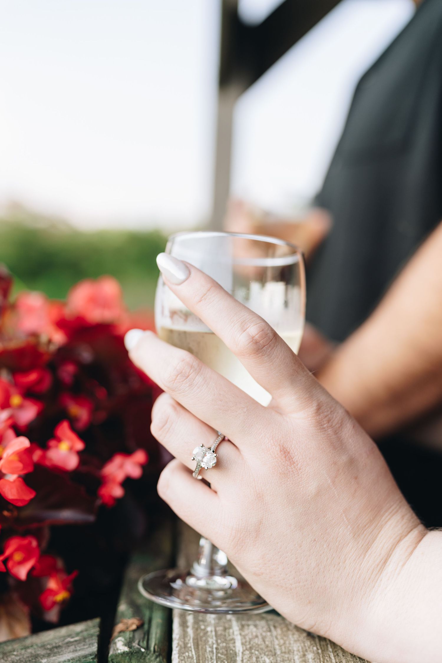 Close-up of an engagement ring on a woman's hand holding a wine glass near red flowers.