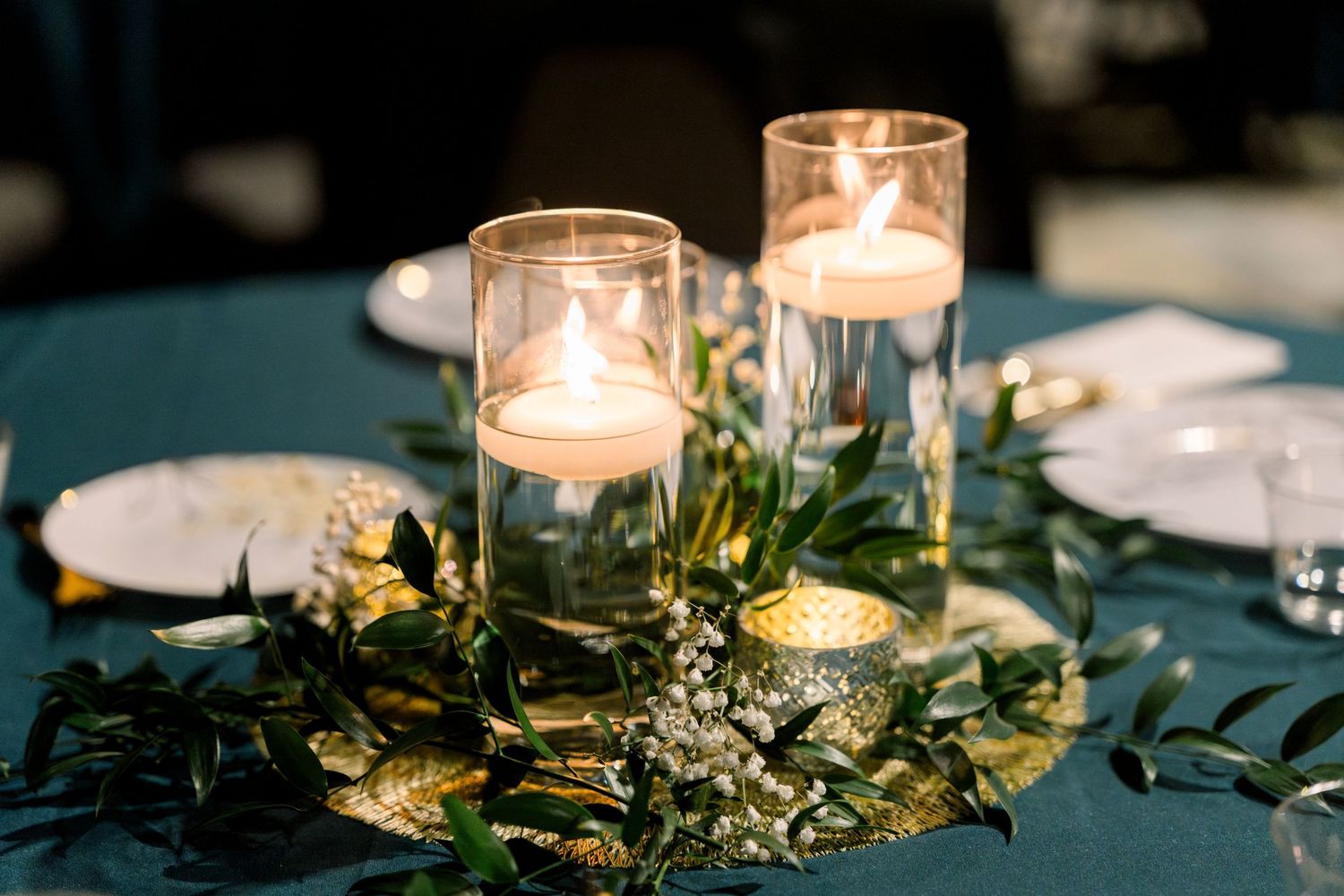 Elegant wedding centerpiece with floating candles in glass cylinders on gold charger with green garland.