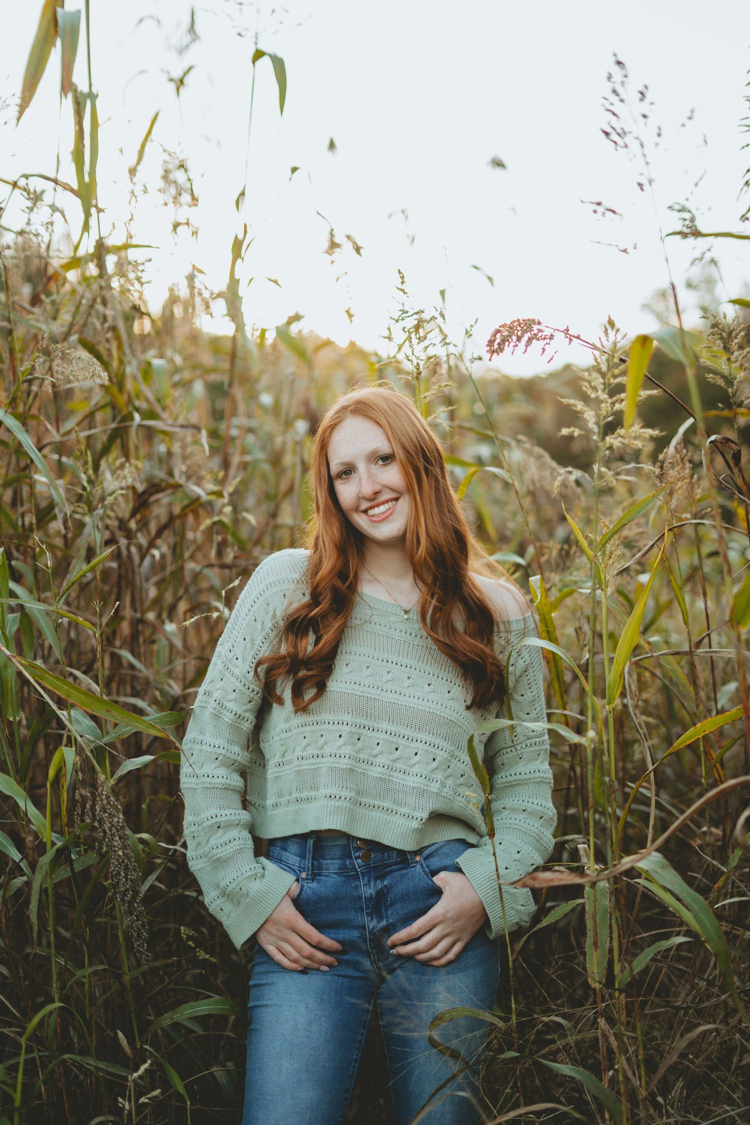 Smiling redhead girl in mint sweater and jeans posing among tall autumn grasses.