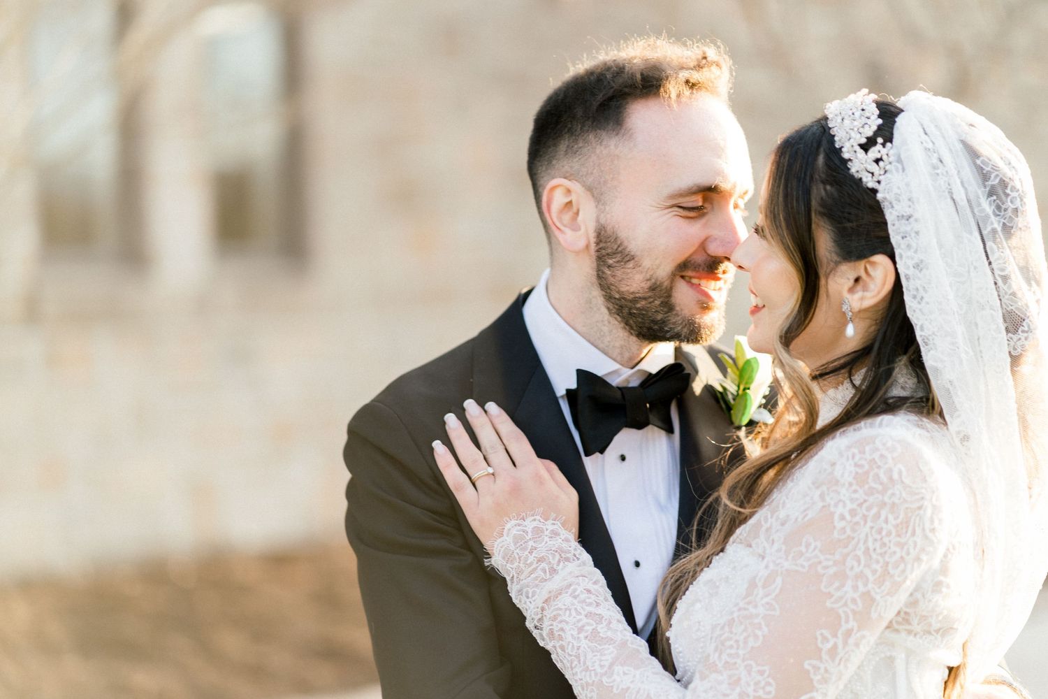 Bride in lace veil and groom in black tuxedo share intimate moment near stone building in warm sunlight.