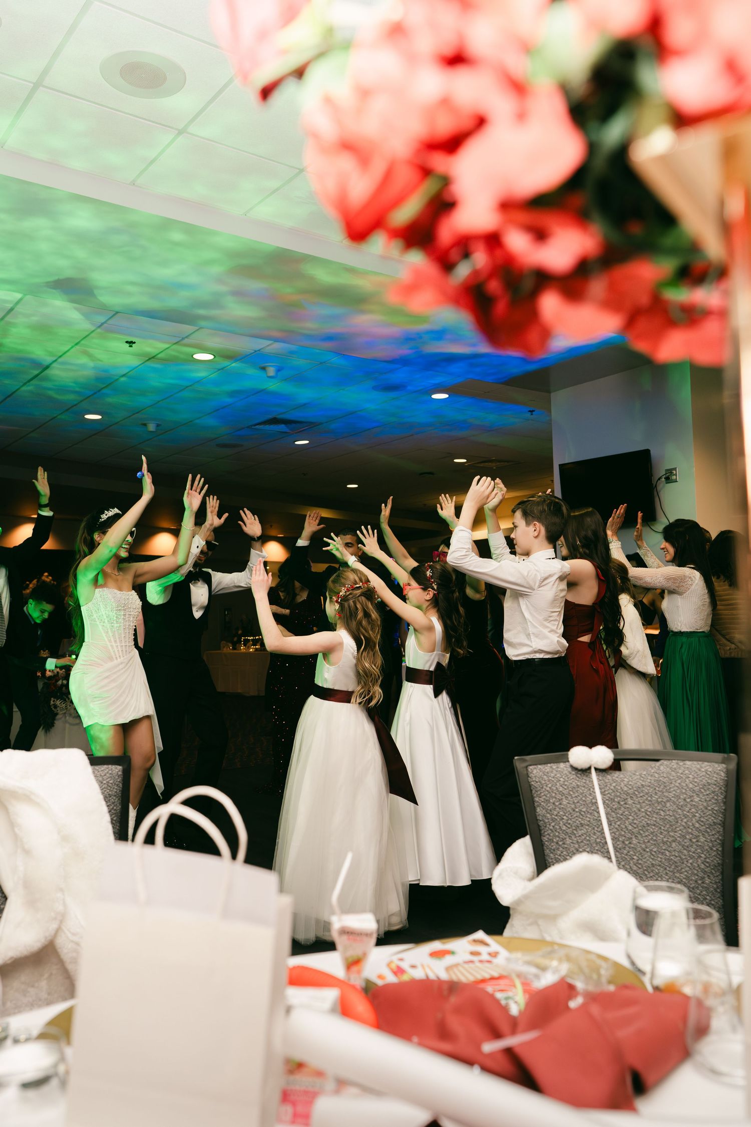Wedding guests raise hands on dance floor with colorful lighting during lively wedding reception celebration.