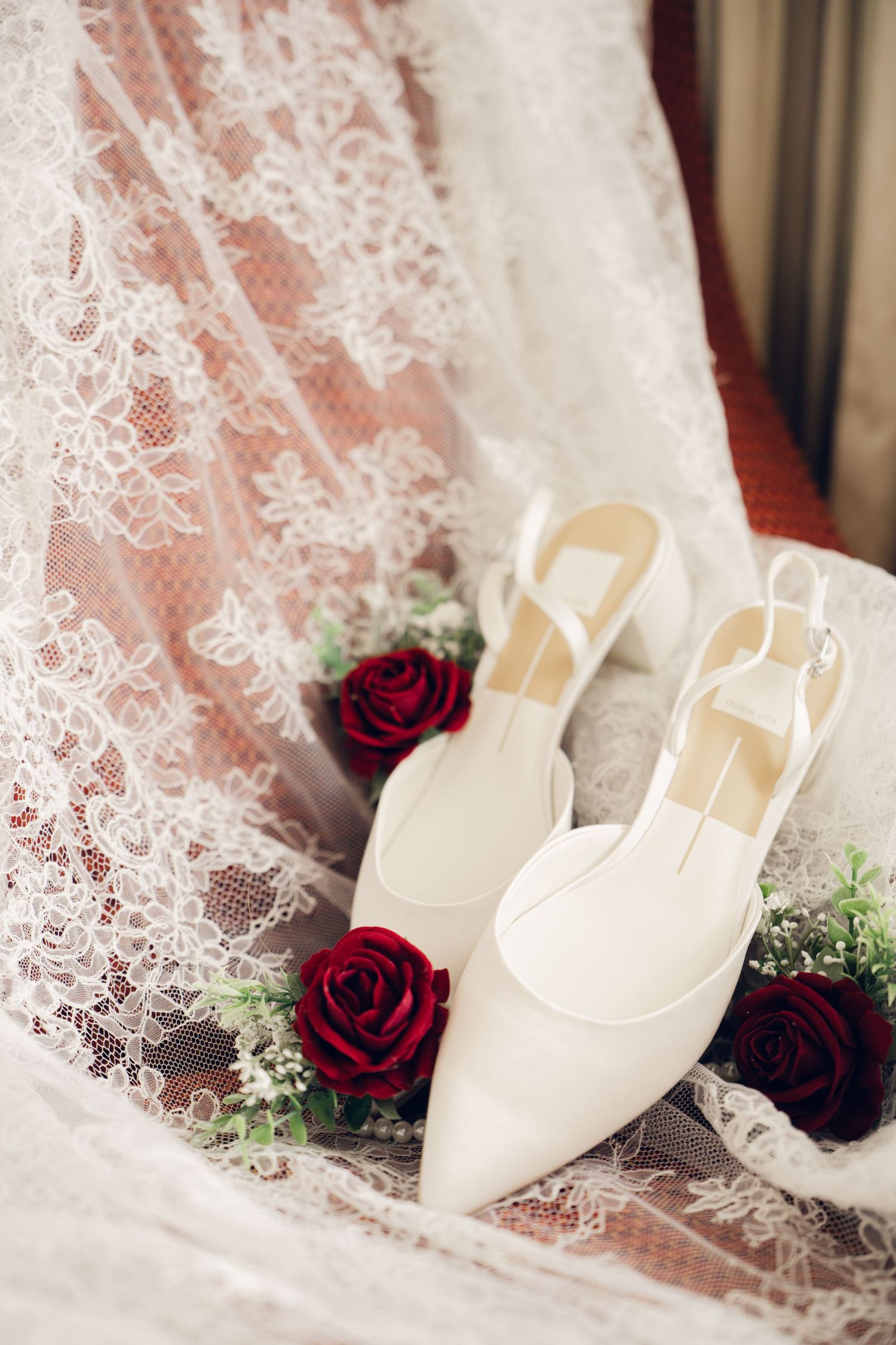 White pointed-toe bridal heels with red rose boutonnieres resting on an intricate lace wedding dress.