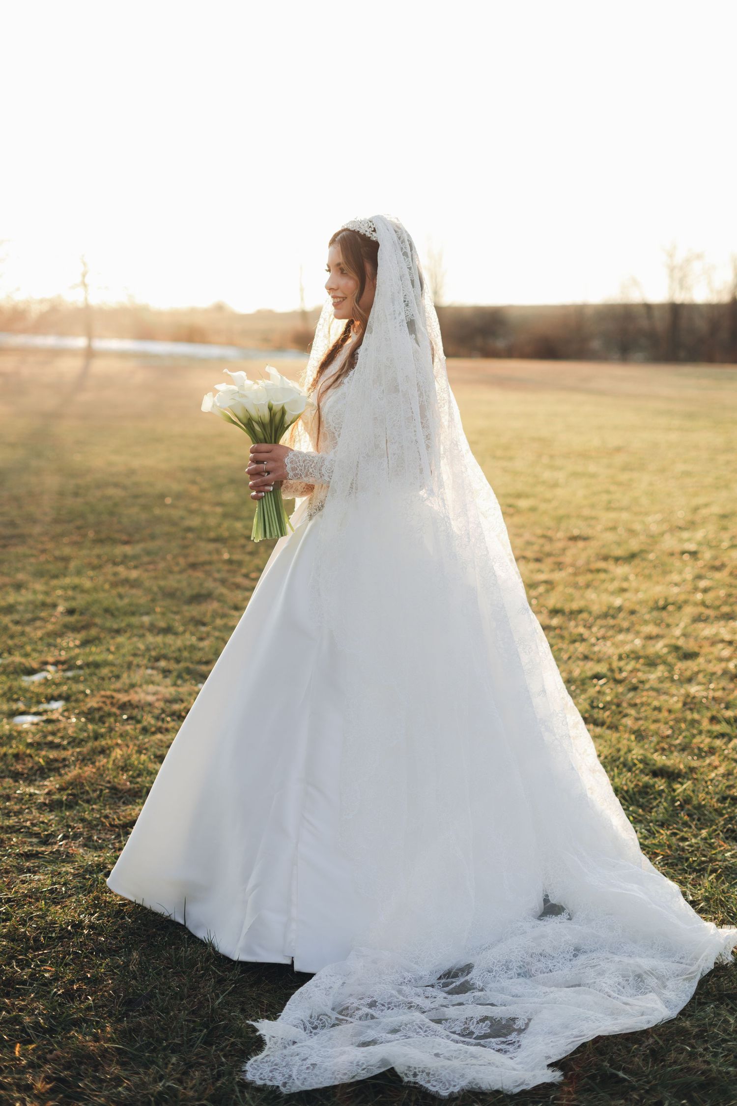 Bride in flowing lace wedding gown and veil holding calla lily bouquet in golden sunset field.