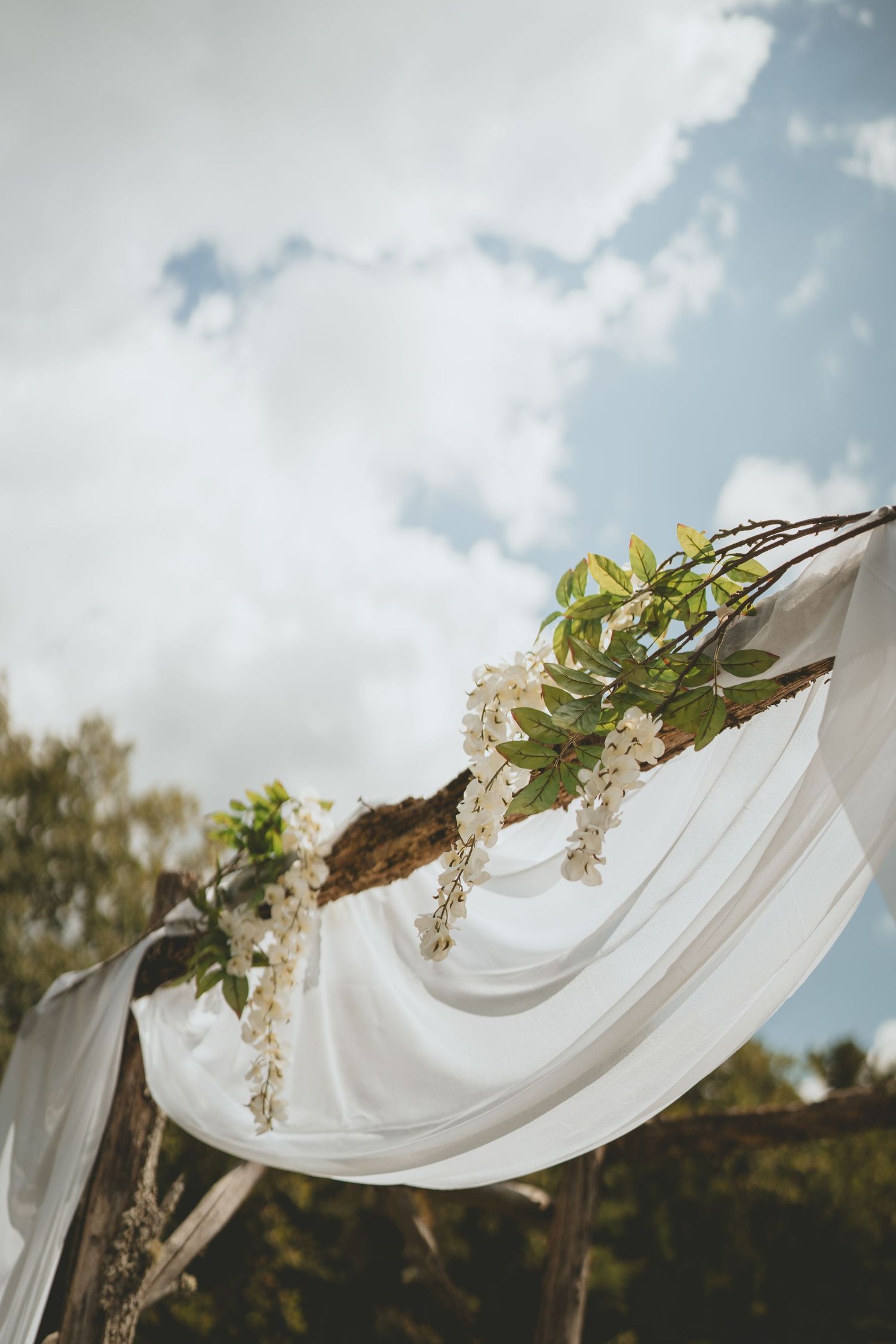 Rustic wooden wedding arch decorated with white wisteria blooms and flowing white fabric under a cloudy sky.
