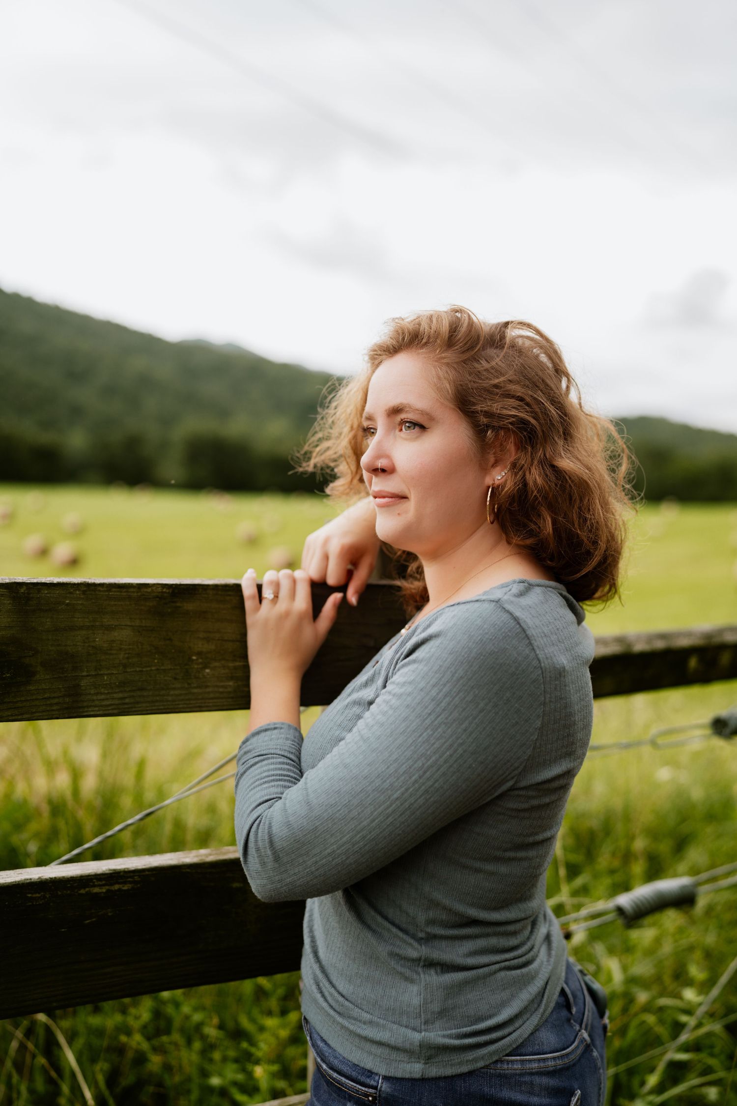Woman in gray long-sleeve top leans on wooden fence gazing at green mountain valley landscape.