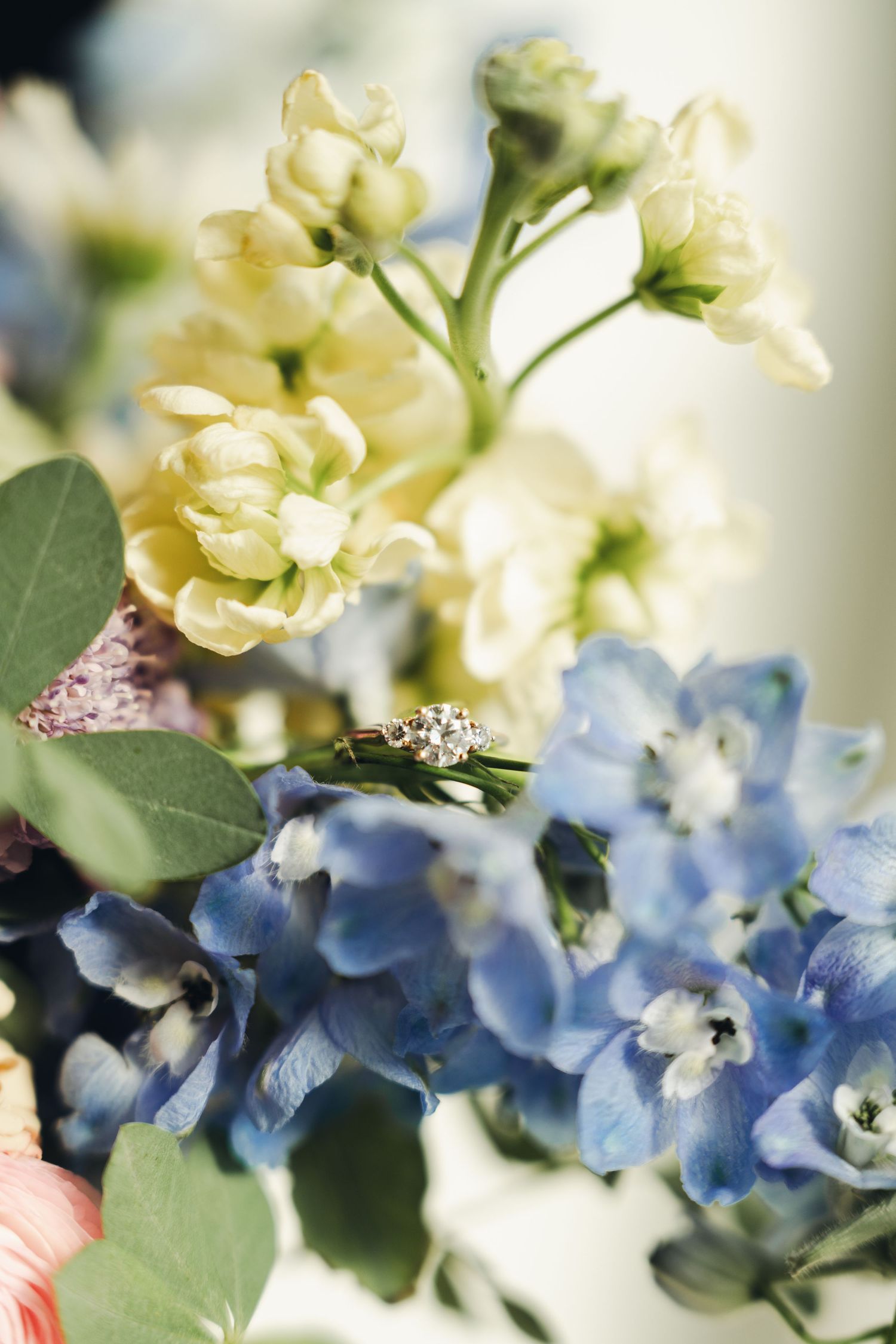 Diamond engagement ring resting on blue and white wedding bouquet with delphinium and stock flowers.