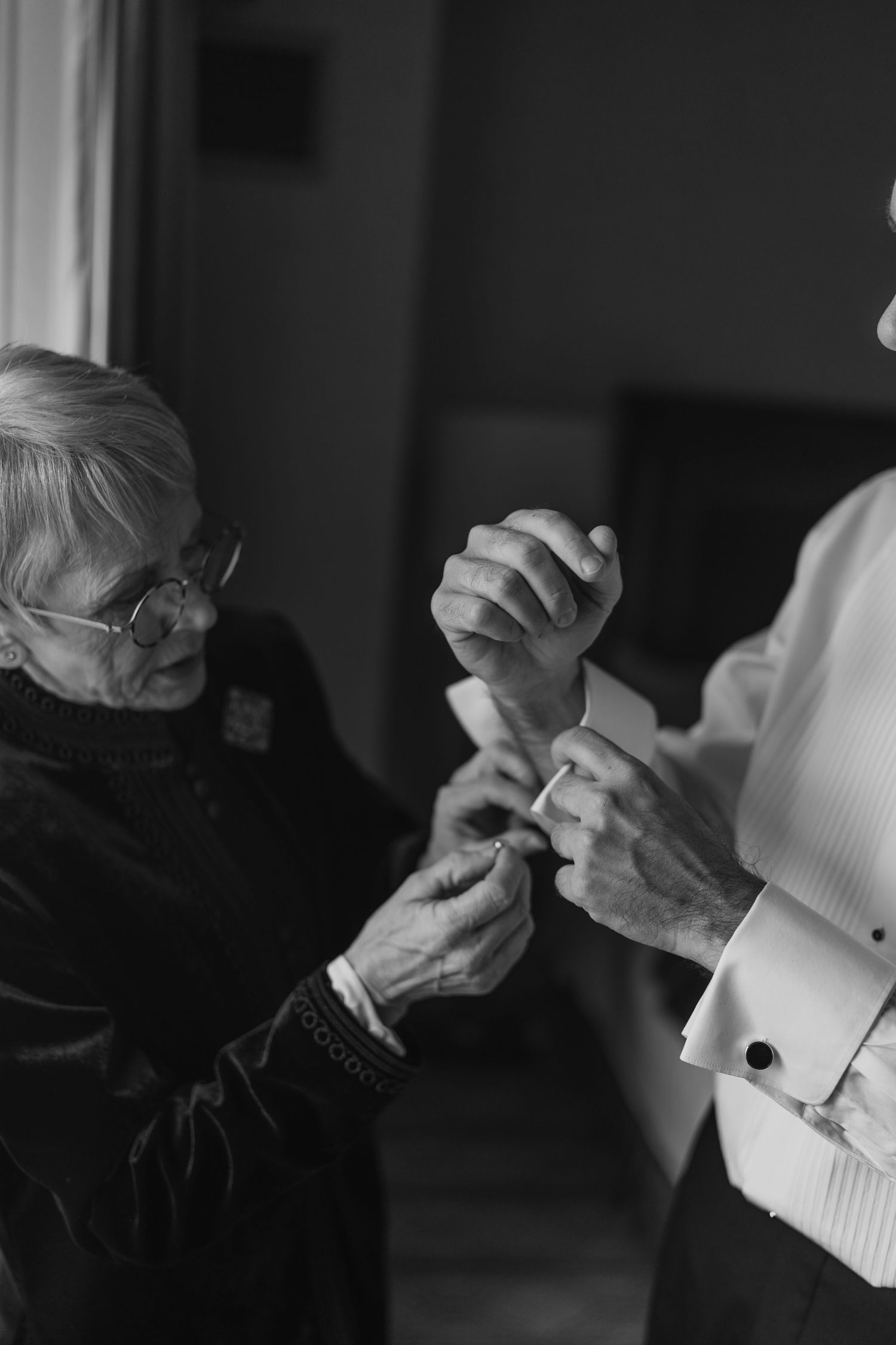 Black and white photo of elderly woman helping groom fasten cufflinks before wedding ceremony.