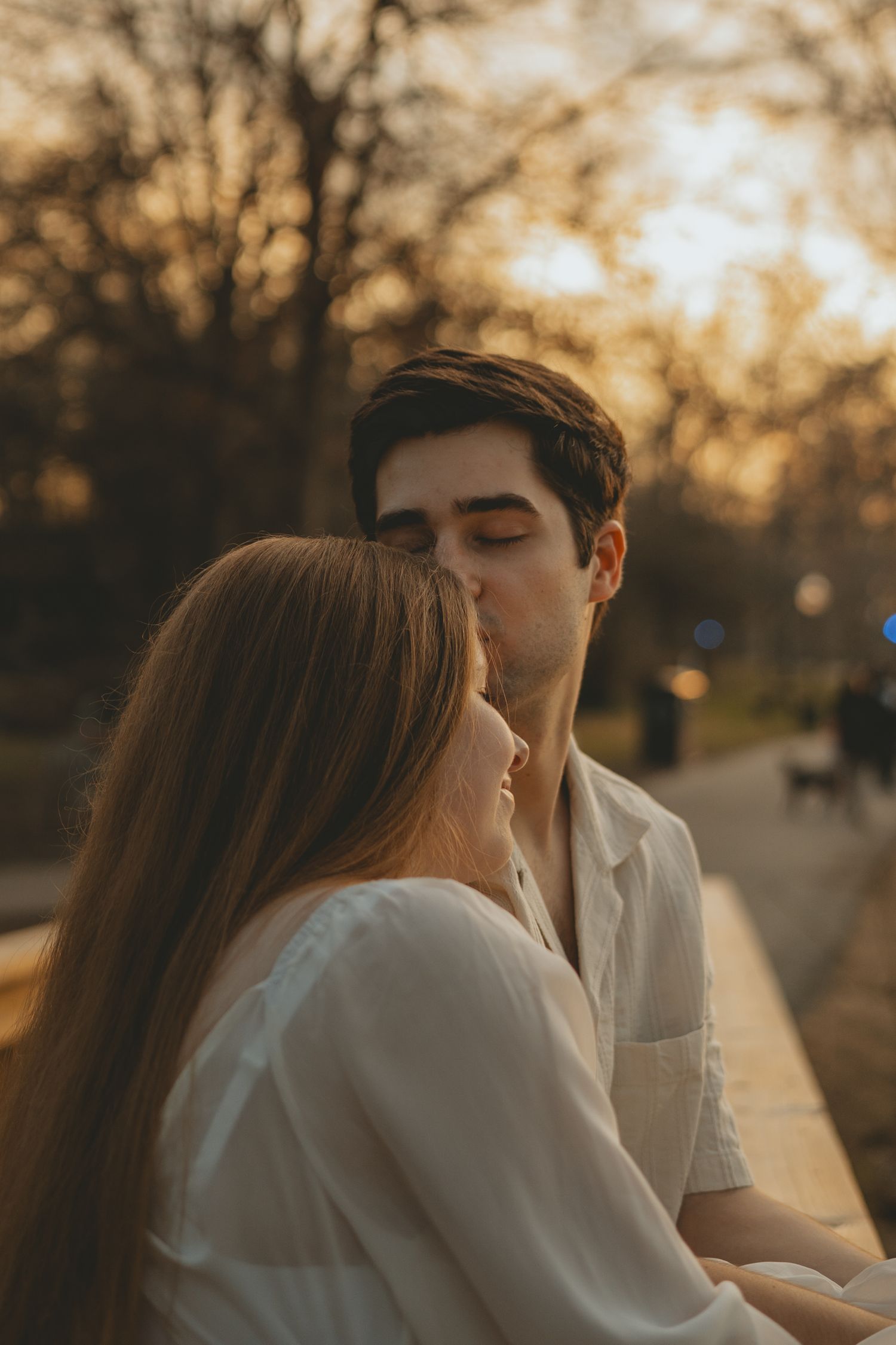 A man gently kisses a woman on the head while sitting together outdoors at warm golden hour.