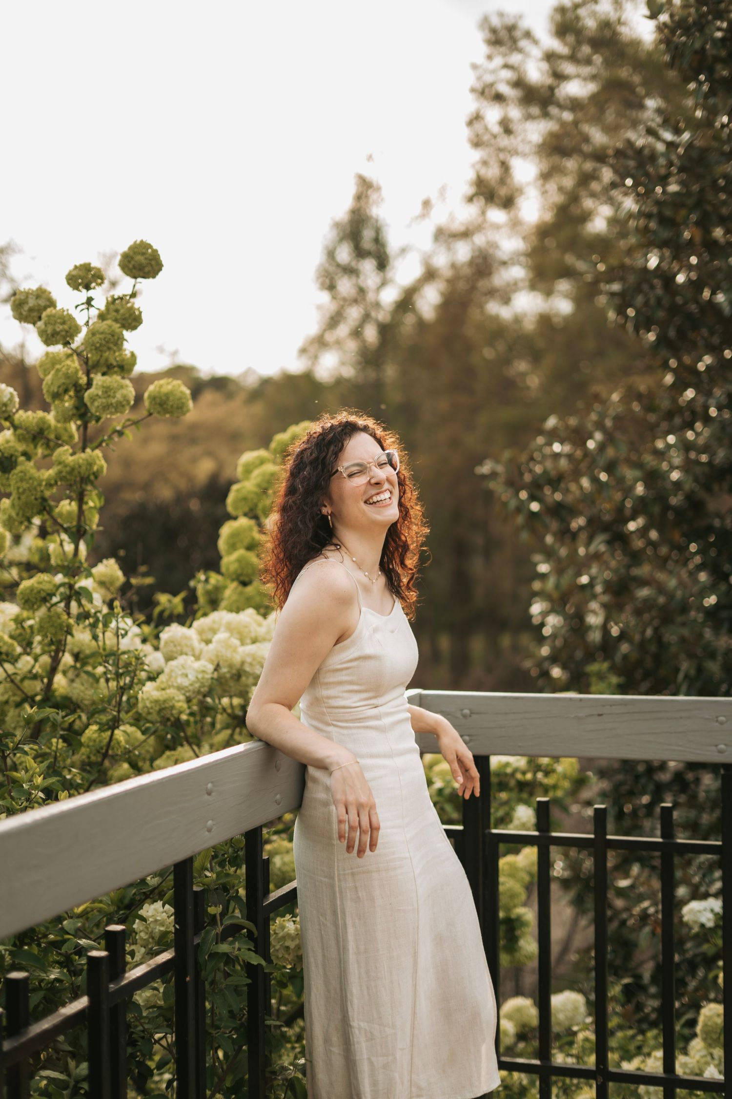A smiling woman with curly red hair in a white dress leans on a railing surrounded by green hydrangeas.