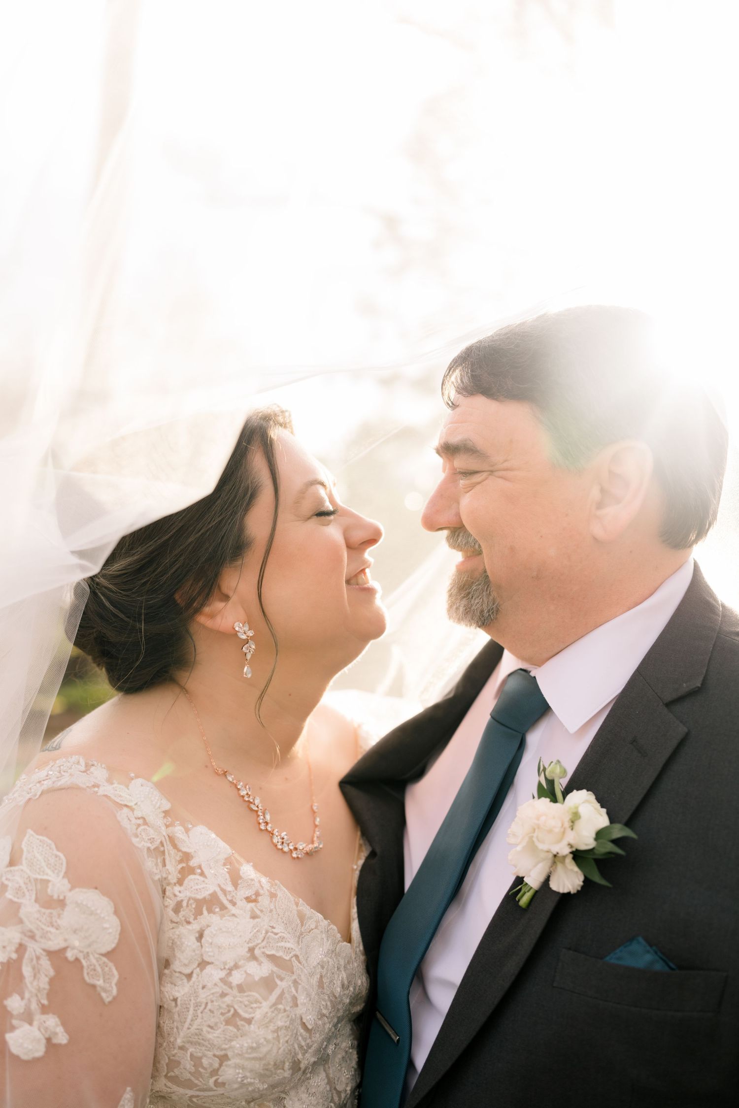 Bride and groom sharing an intimate moment under a flowing veil with soft backlit sunlight on their wedding day.