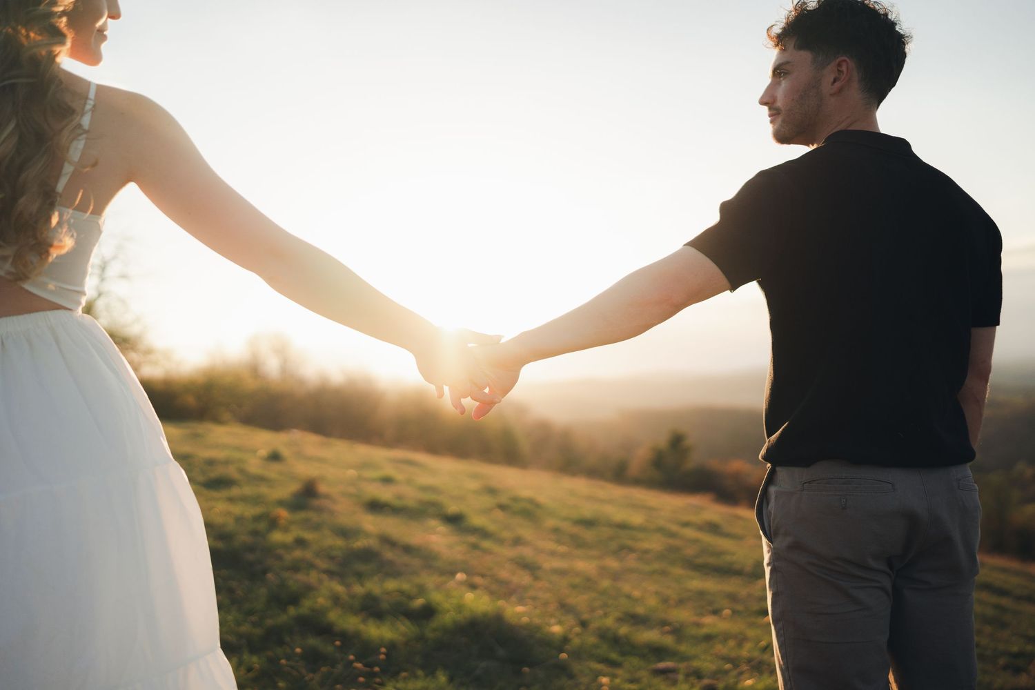 Couple holding hands walking through a sunlit green meadow at golden hour sunset.