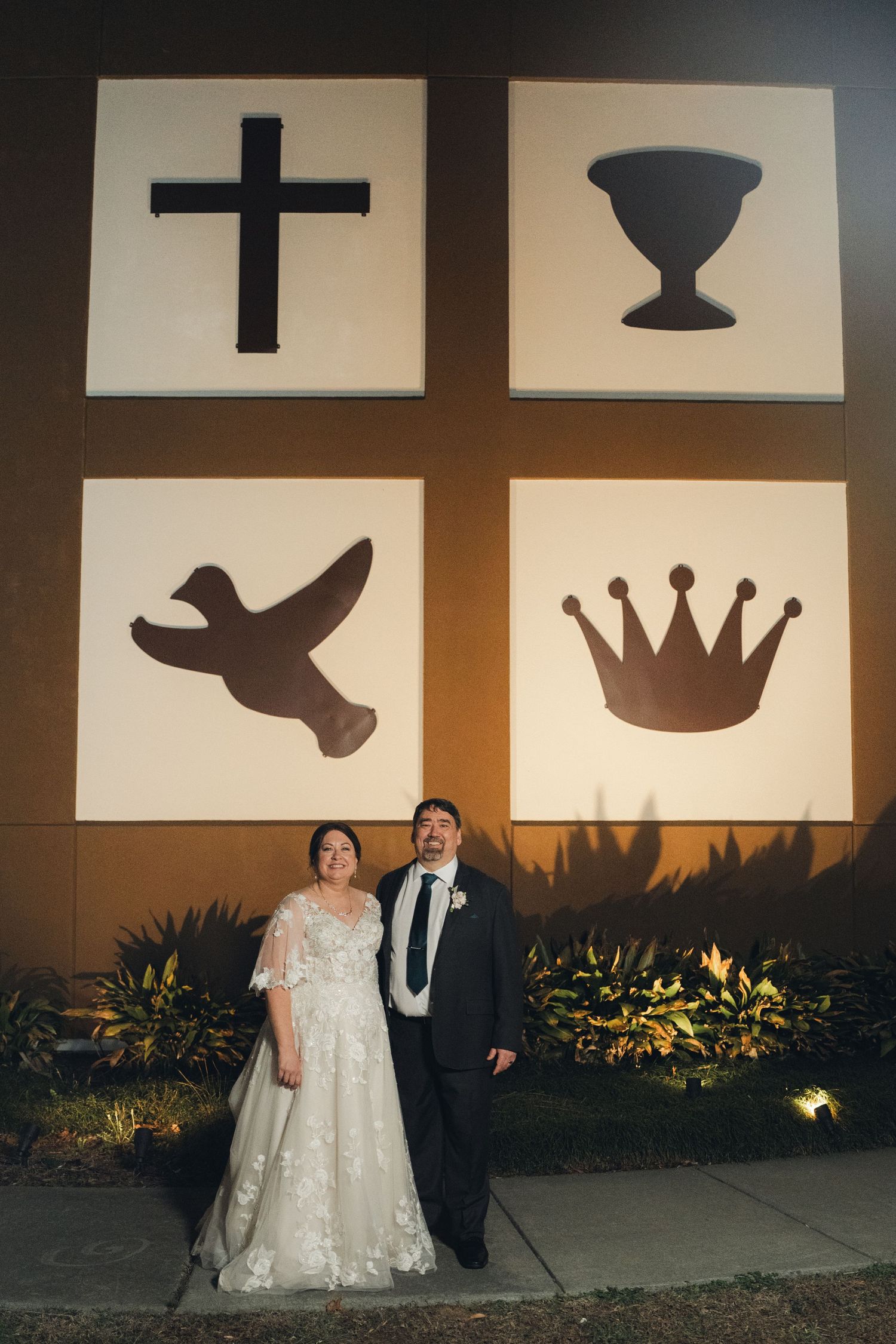 Bride and groom pose at night beneath illuminated Christian symbols on church exterior wall after wedding.
