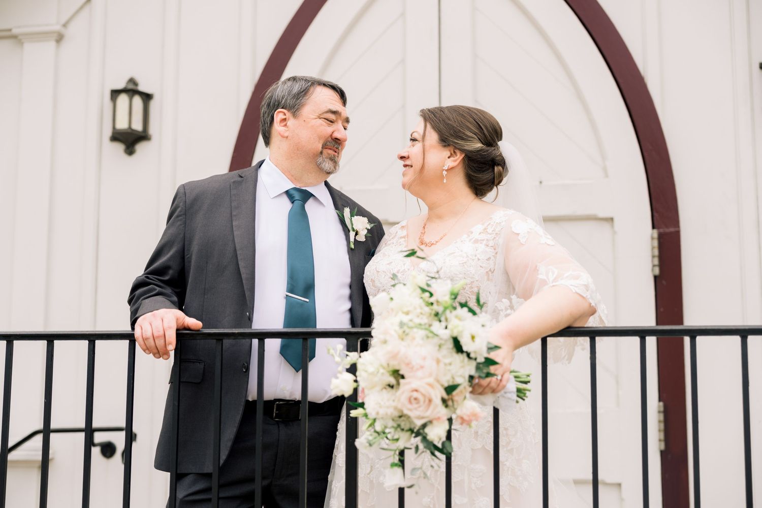 Bride and groom laughing together on a balcony in front of a white church entrance.