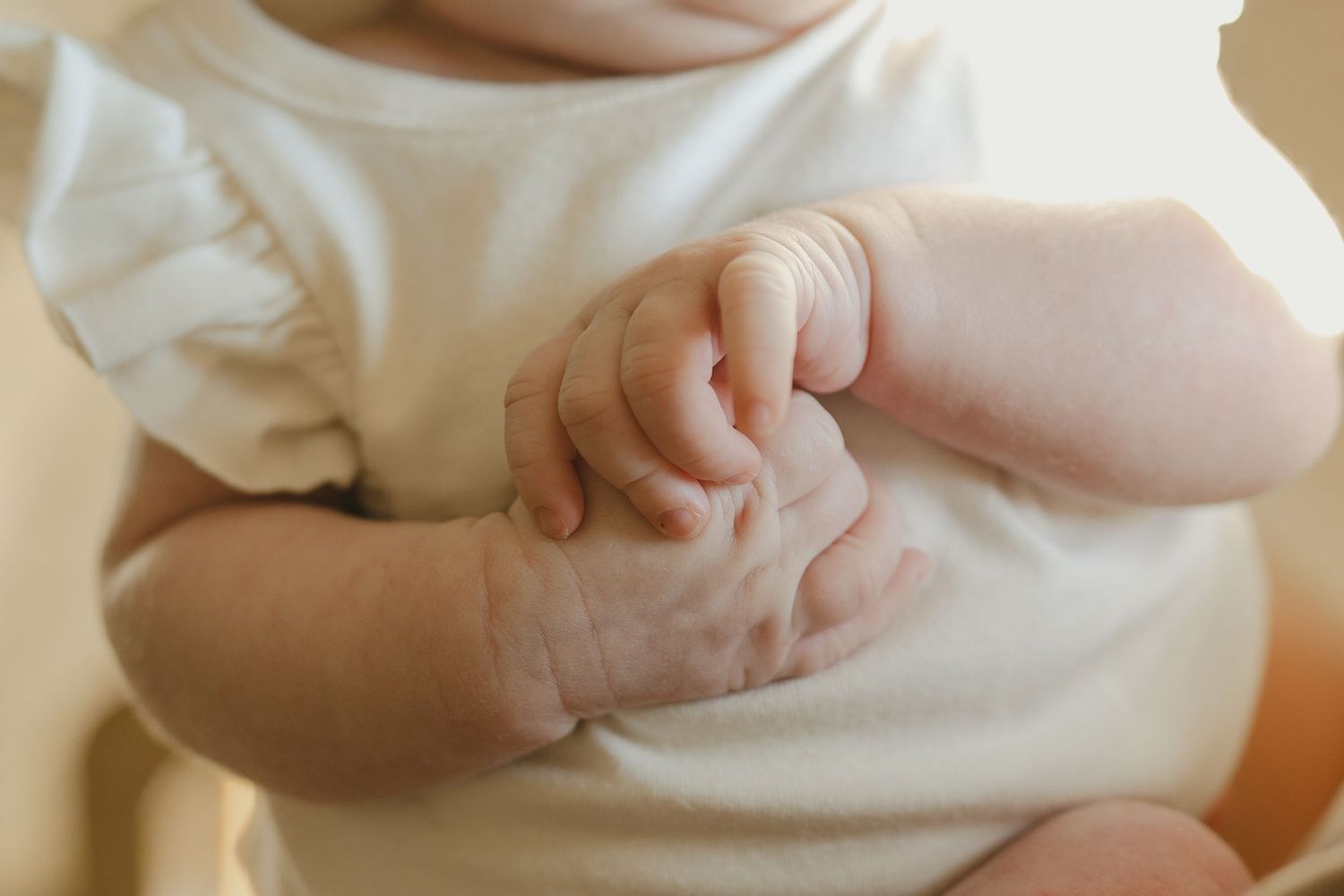 Close-up of newborn baby girl in white ruffle onesie with tiny hands clasped together.