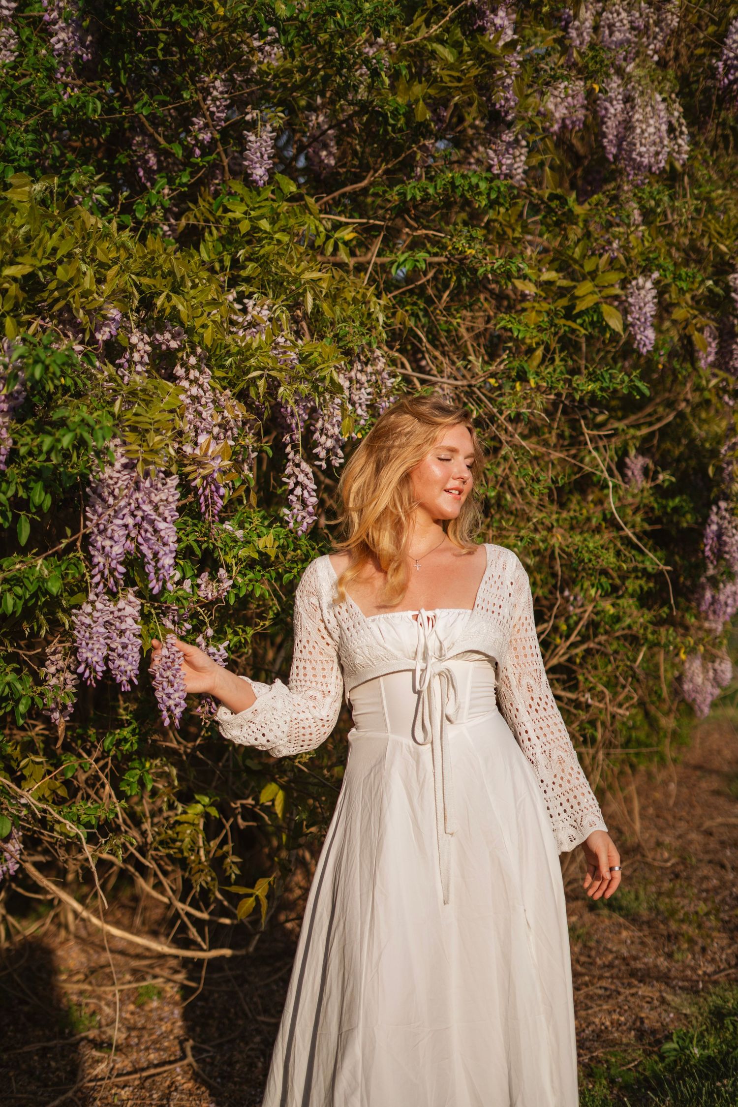 A blonde woman in a white crochet dress reaches toward purple wisteria flowers in a garden.