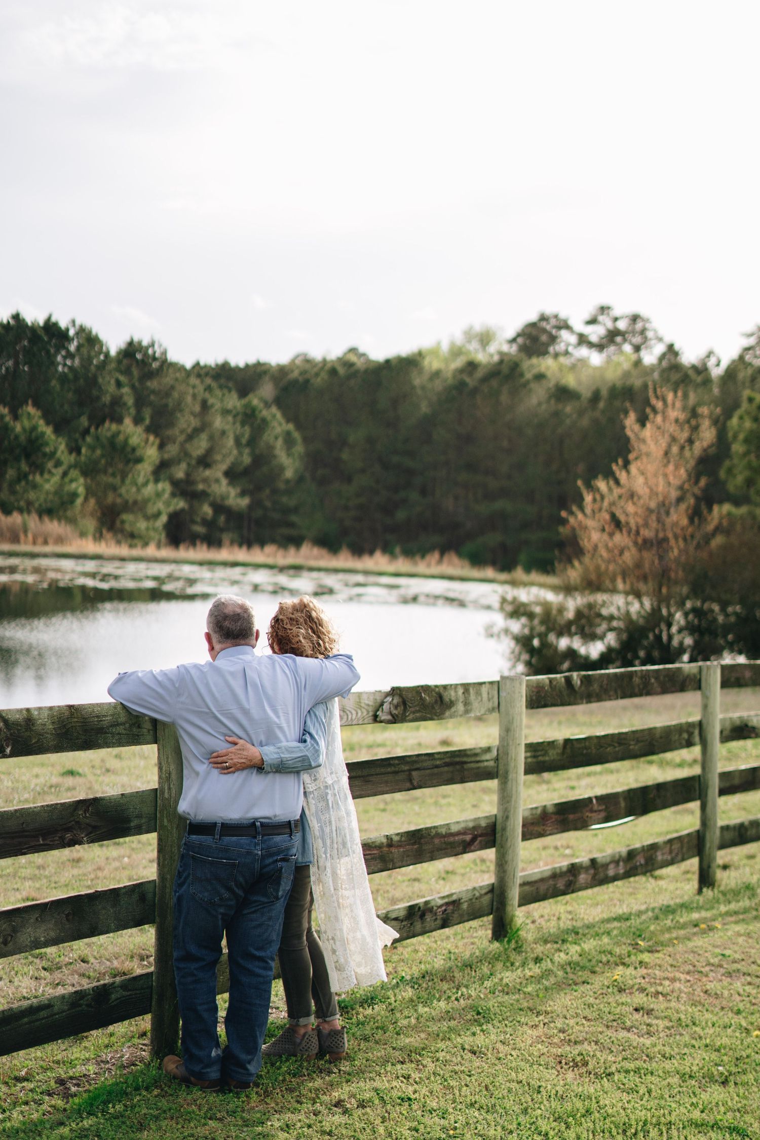Couple embracing while looking at peaceful pond from wooden farm fence in rural setting.