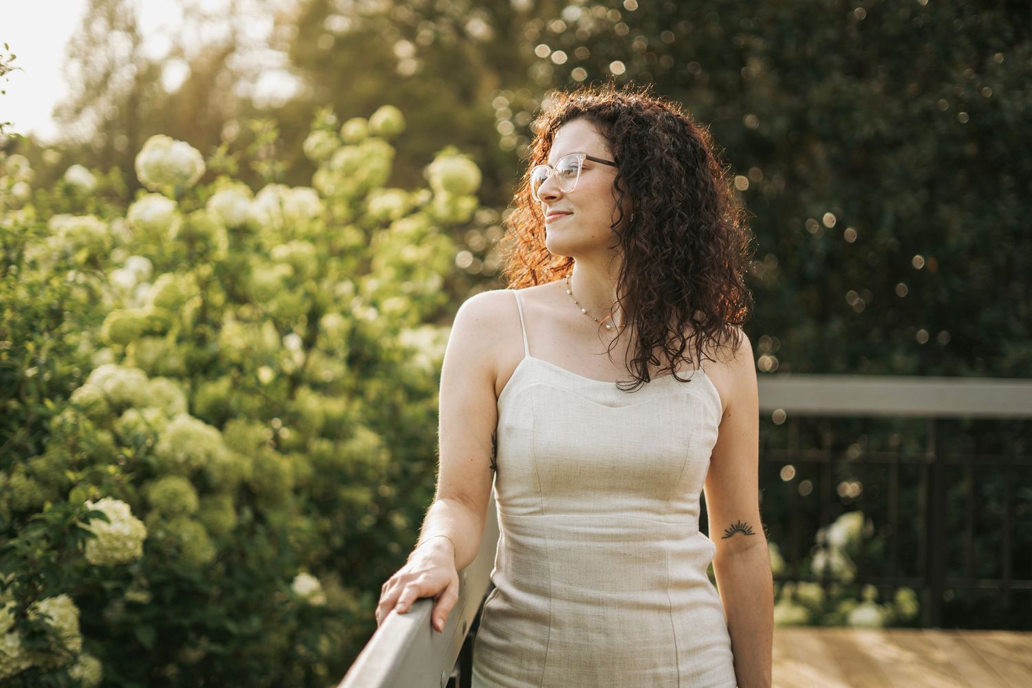 Curly-haired woman in beige dress smiling outdoors near white hydrangea bushes in sunlight.