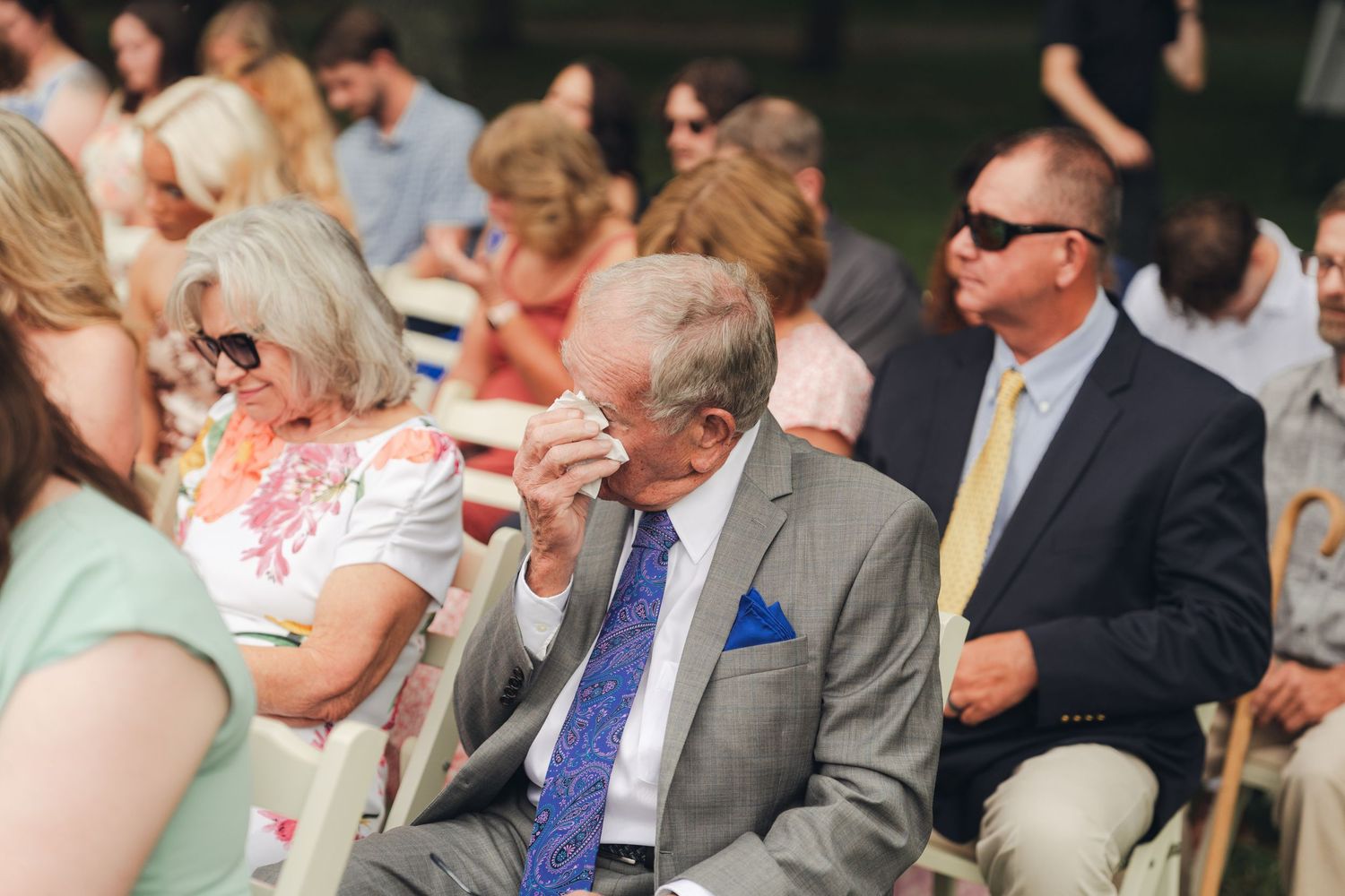 Elderly man wiping tears at an outdoor wedding ceremony, surrounded by guests in formal attire.