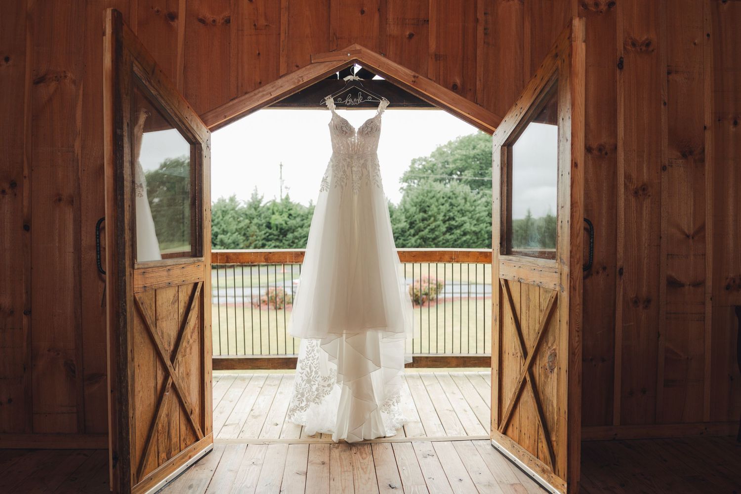 Elegant white wedding dress hanging in rustic barn doorway with wooden doors open to lush green outdoor scenery.