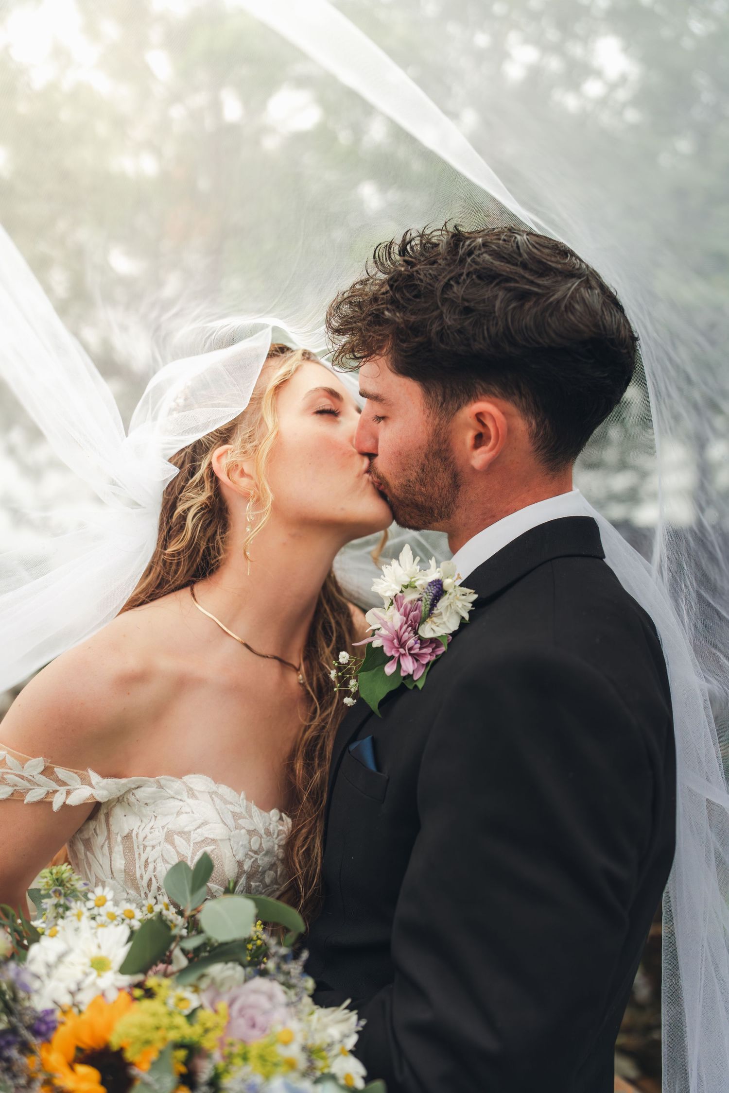 Bride and groom sharing a romantic kiss under a flowing veil with a wildflower bouquet.