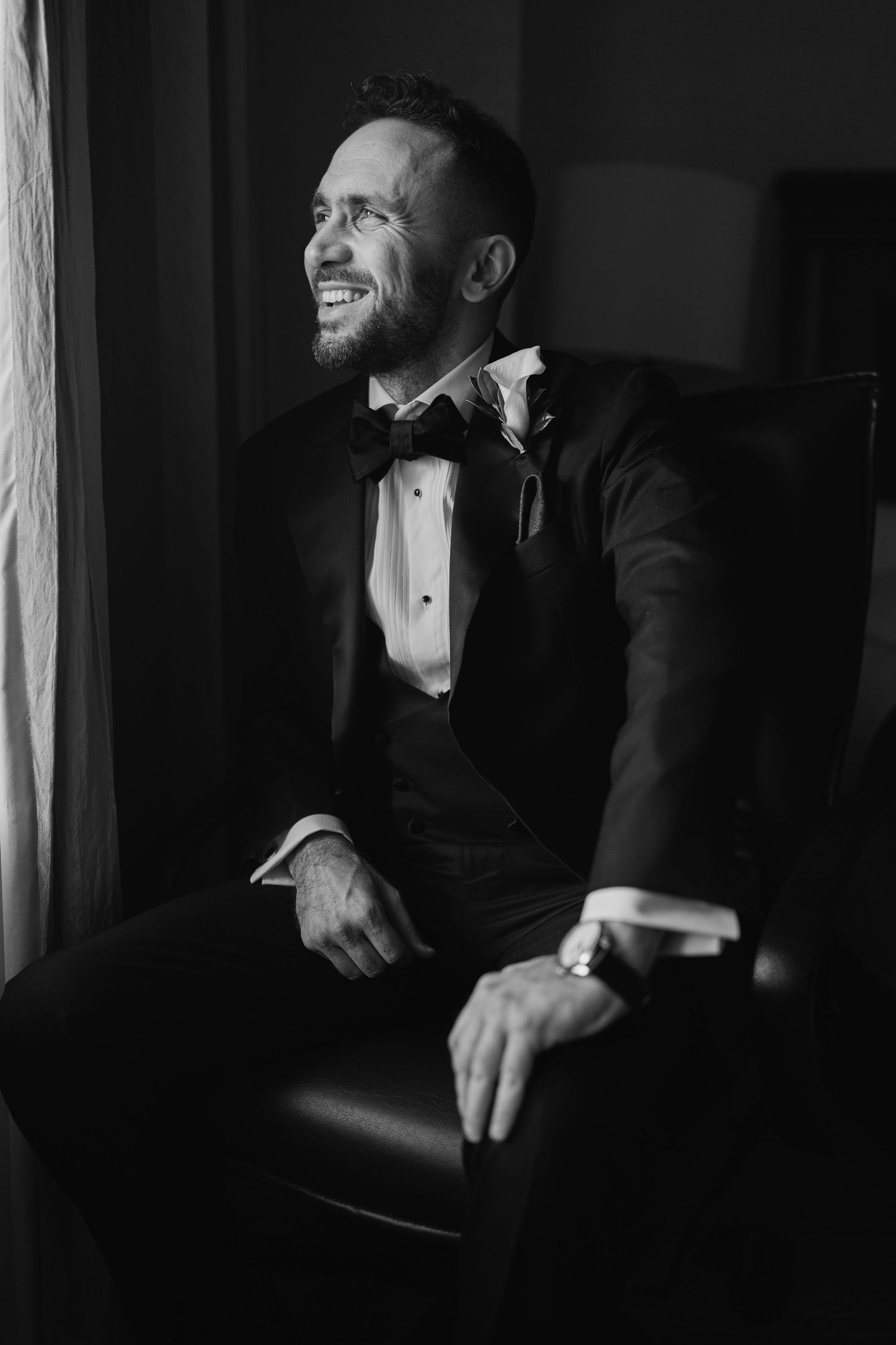 Happy groom in tuxedo sitting in chair by window, black and white.