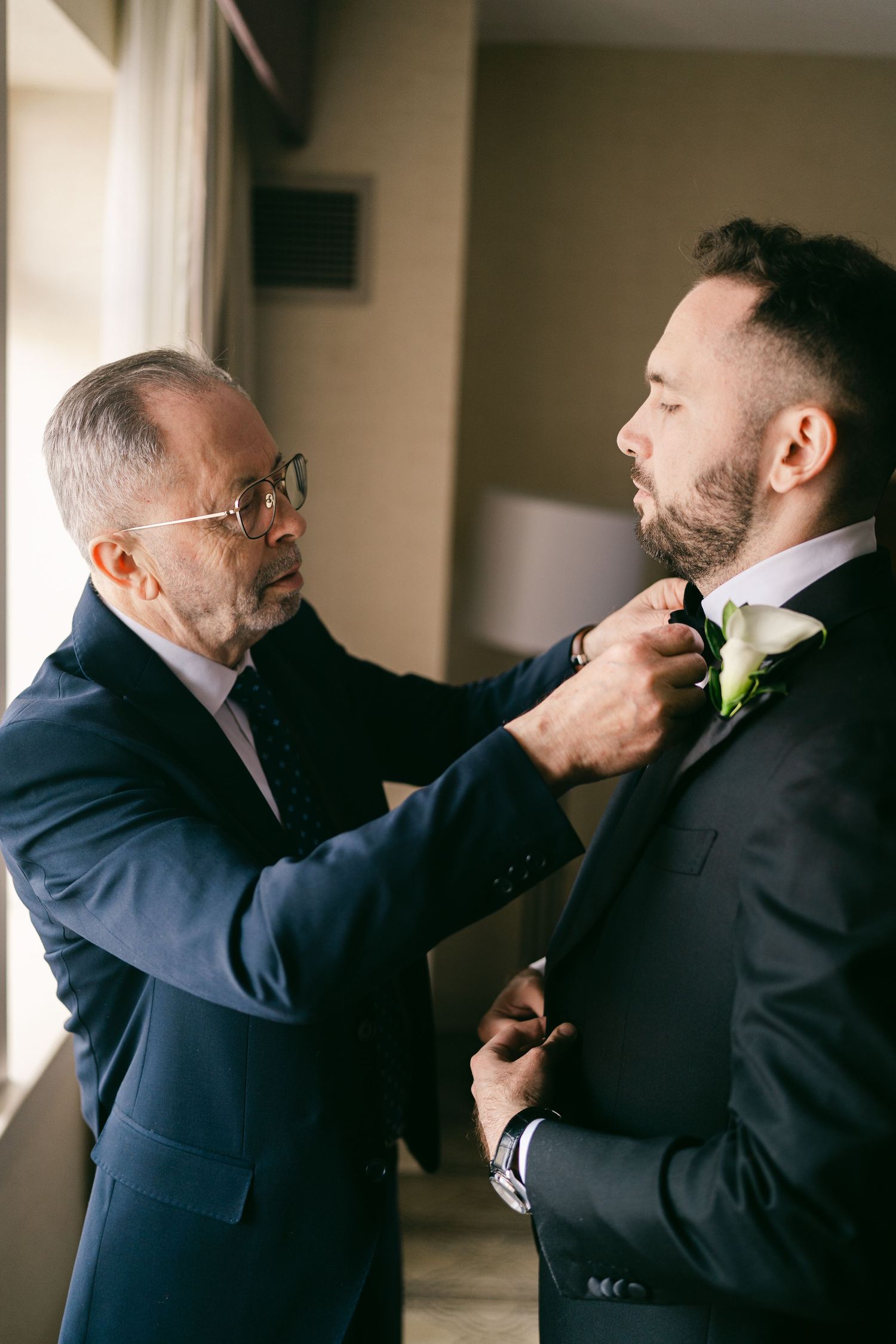 Older man helping groom adjust tie before wedding ceremony indoors.