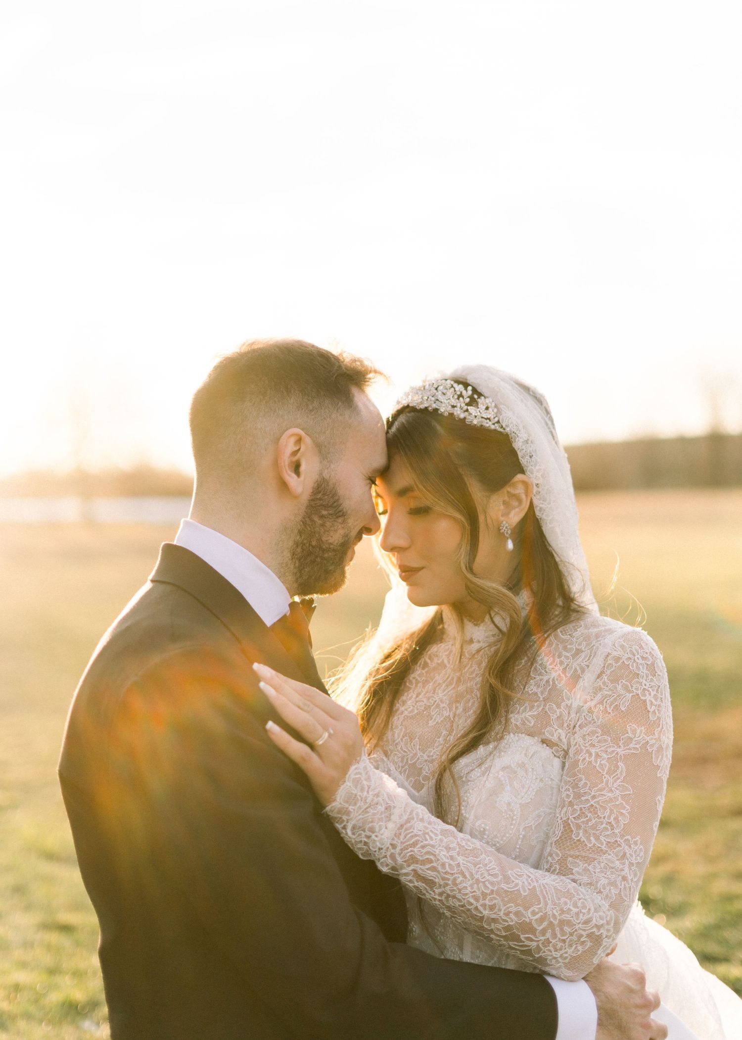 Bride and groom share romantic forehead-to-forehead moment bathed in golden sunset light in open field.