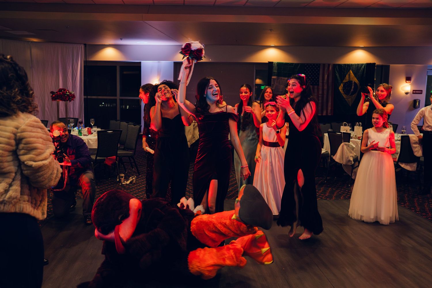 Excited guests reach for bouquet toss as bride throws flowers at colorful wedding reception celebration.