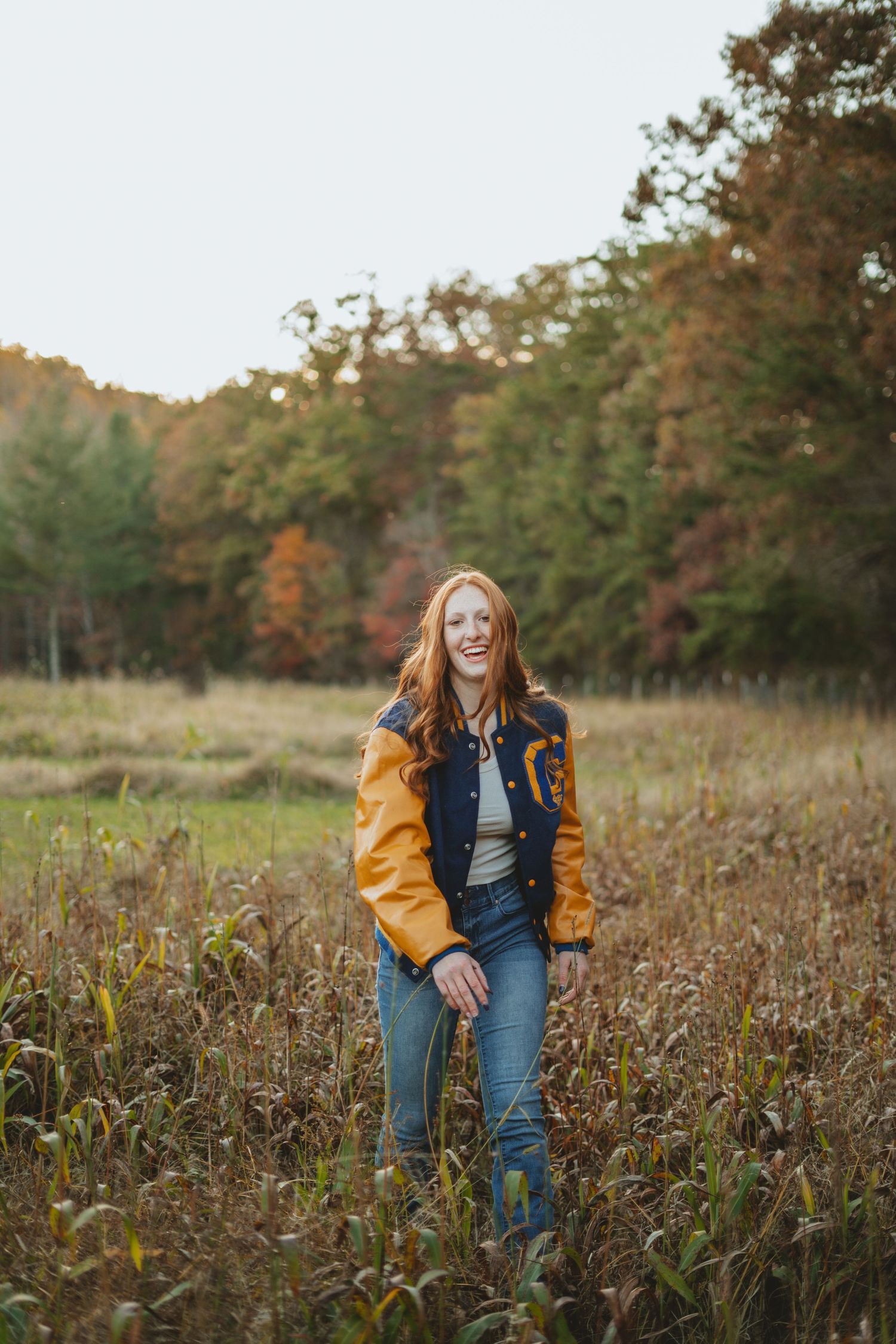Laughing redhead girl in varsity jacket standing in golden autumn field at sunset.